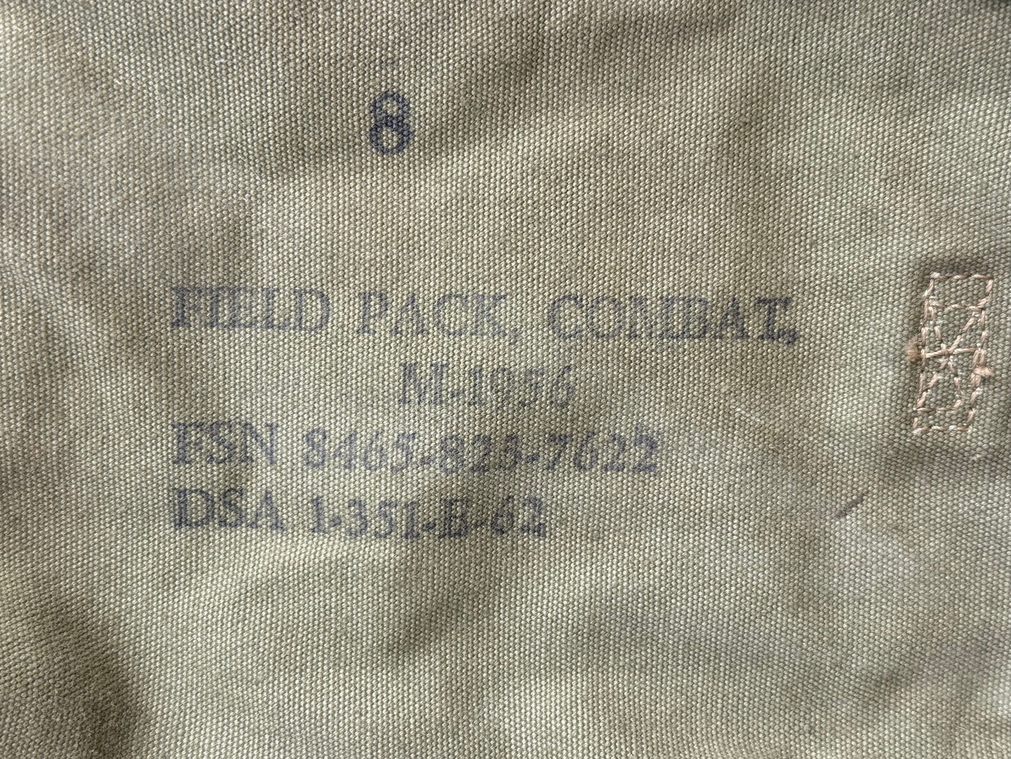 Close-up of a beige or khaki fabric surface with printed dark gray text reading 'FIELD PACK, COMBAT,' along with a number 8 and a small rectangular woven label or patch on the right.