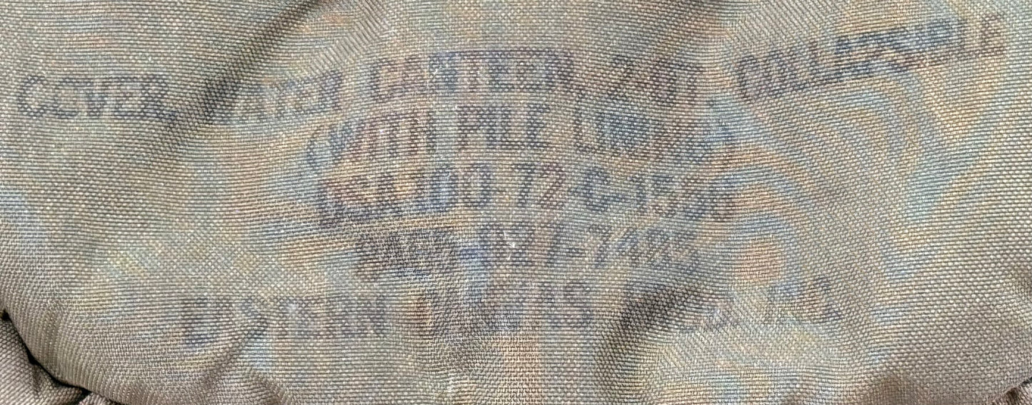 Close-up of a beige canvas cover with blue printed text reading 'COVER WATER CANTEEN 2 QT COLLAPSIBLE (WITH PILE LINING) OSA100-72-C-1596 9406-027-7746 EASTERN CANVAS PROD. INC.