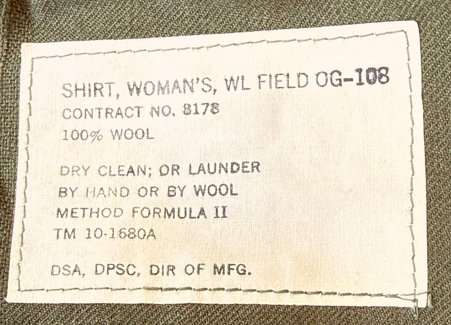 A close-up of a sewn fabric label on an olive green shirt, reading 'SHIRT, WOMAN'S, WL FIELD OG-108' and '100% WOOL'.