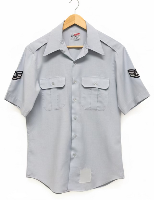 Gray short-sleeve shirt with embroidered logos on a hanger against a yellow wall.