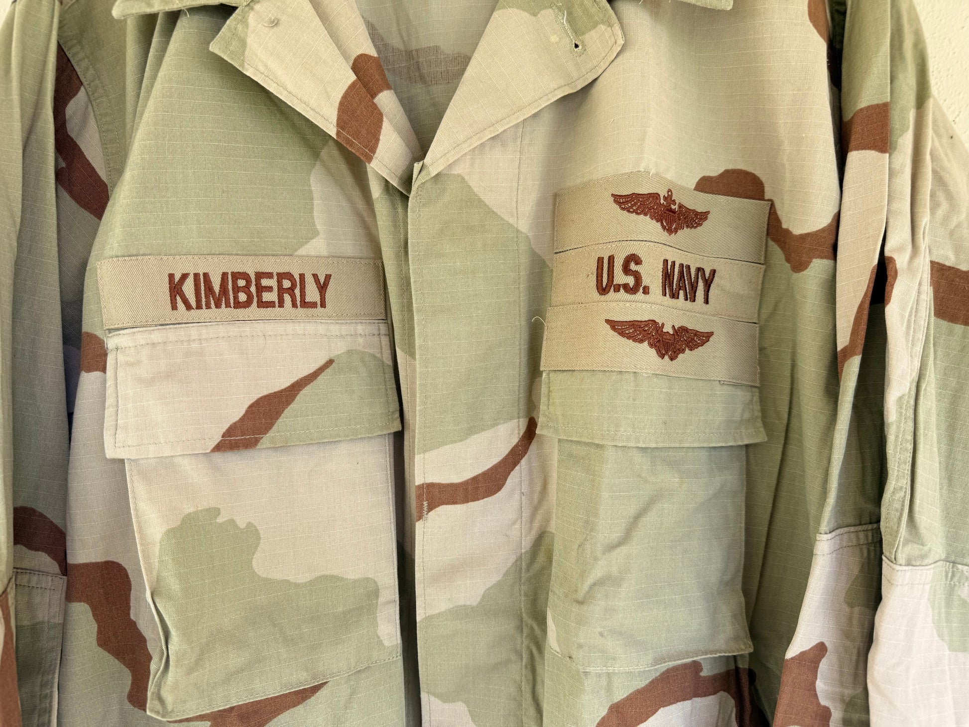 Close-up of a desert camouflage military-style jacket with a flap pocket on the left chest labeled 'KIMBERLY' and a matching pocket on the right chest labeled 'U.S.