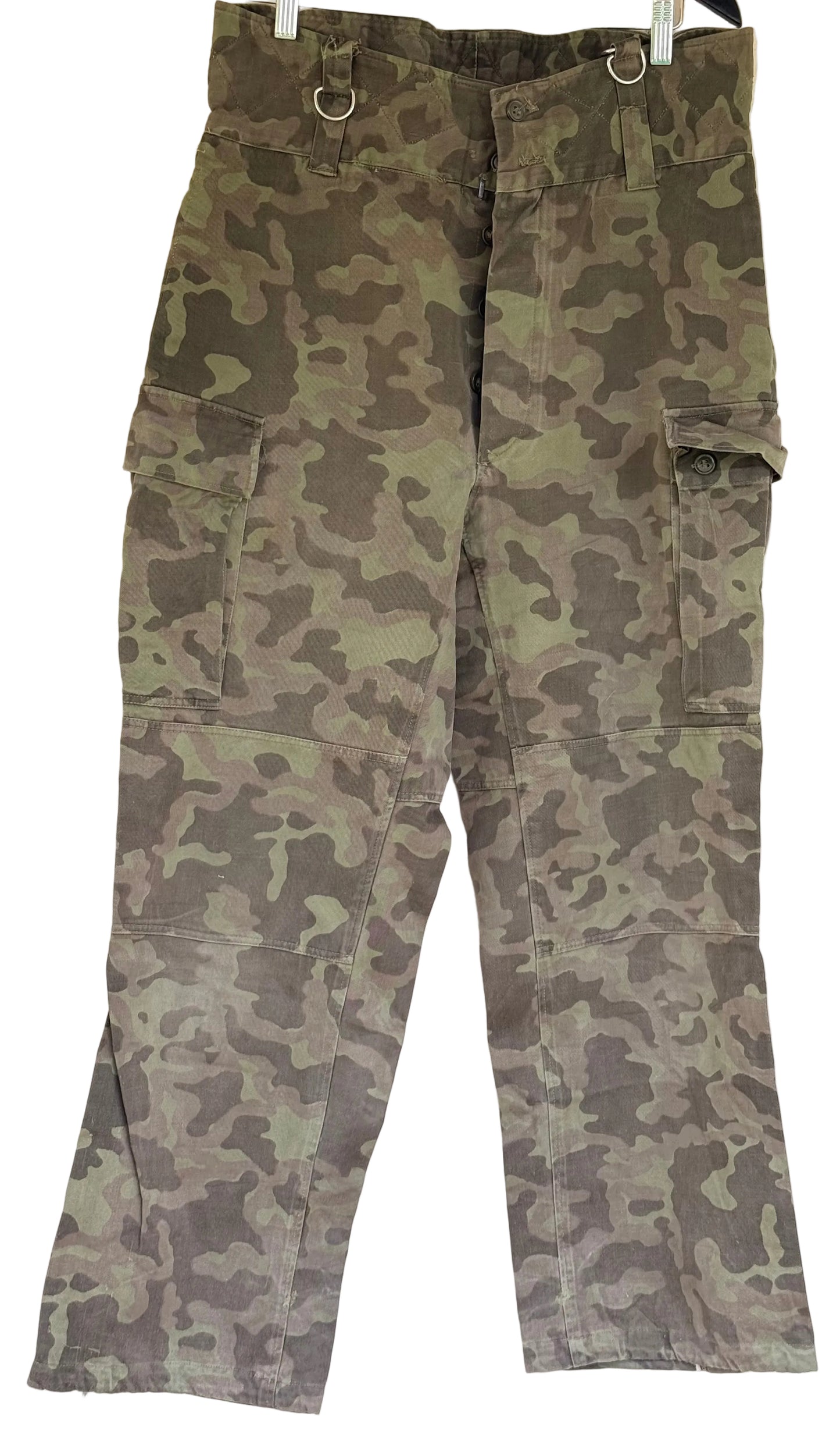 Green and brown camouflage cargo pants hanging on a white wall, featuring multiple utility pockets and metal ring details at the waist.