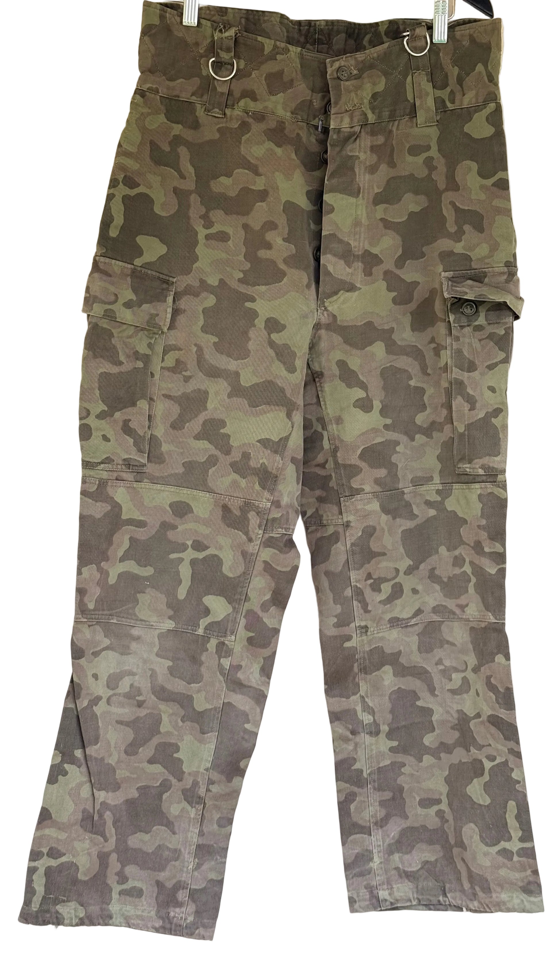 Green and brown camouflage cargo pants hanging on a white wall, featuring multiple utility pockets and metal ring details at the waist.