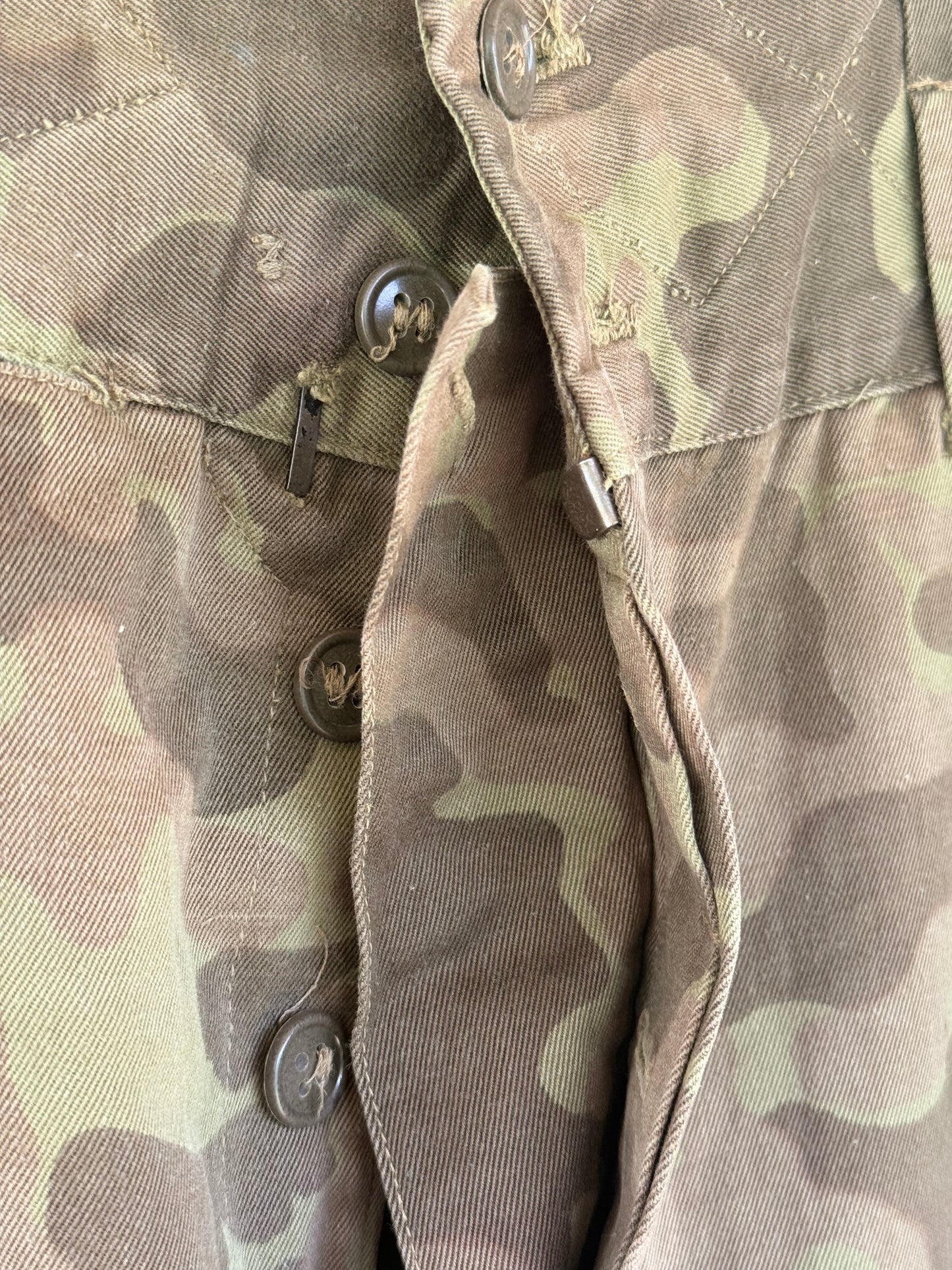 Close-up of a camouflage fabric garment, showing a buttoned pocket flap and the camo pattern in muted green, brown, and tan tones.