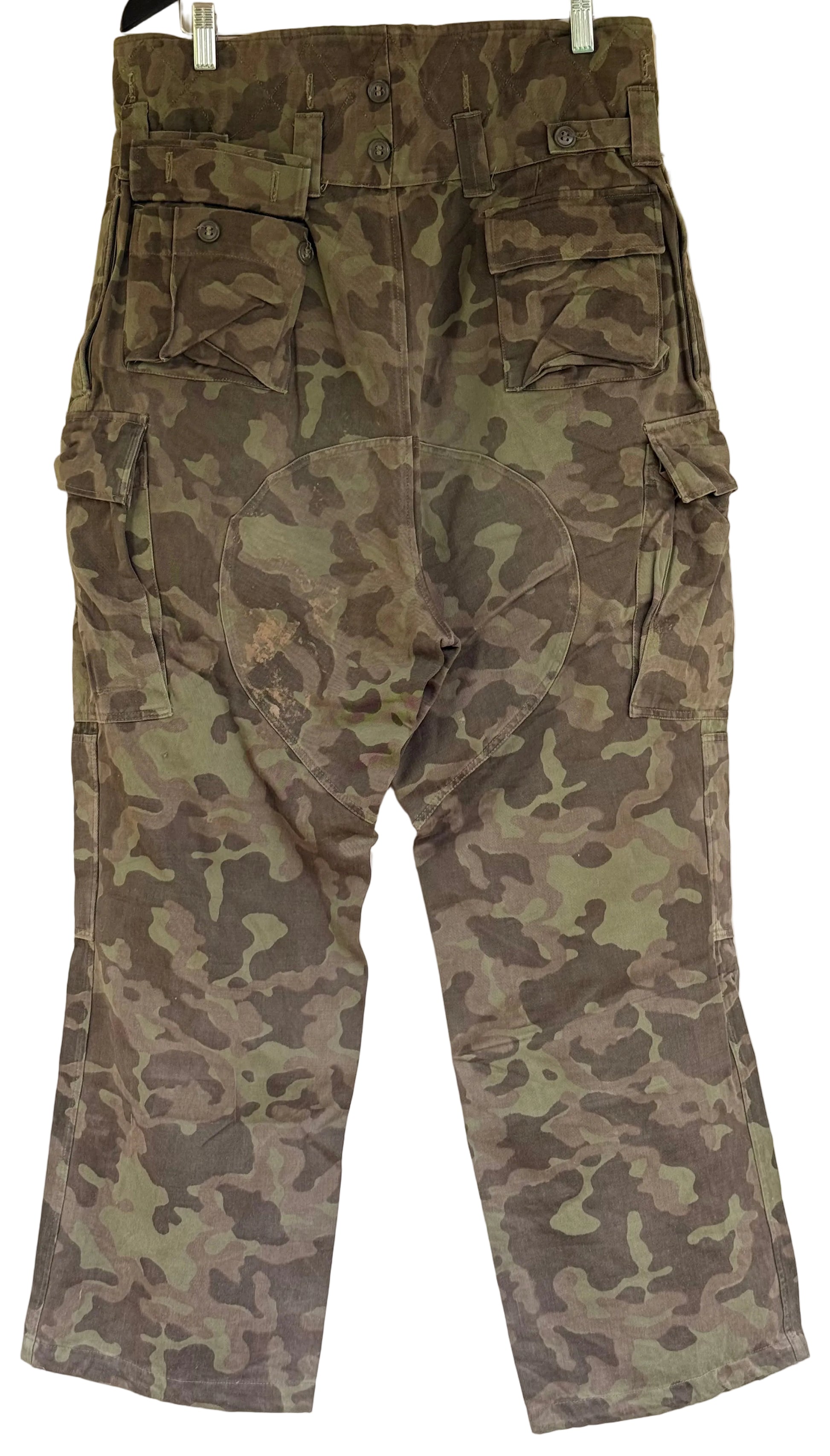 A pair of camouflage cargo pants hanging against a plain light wall, showing multiple flap pockets and a woodland camo pattern in green, brown, and gray tones.