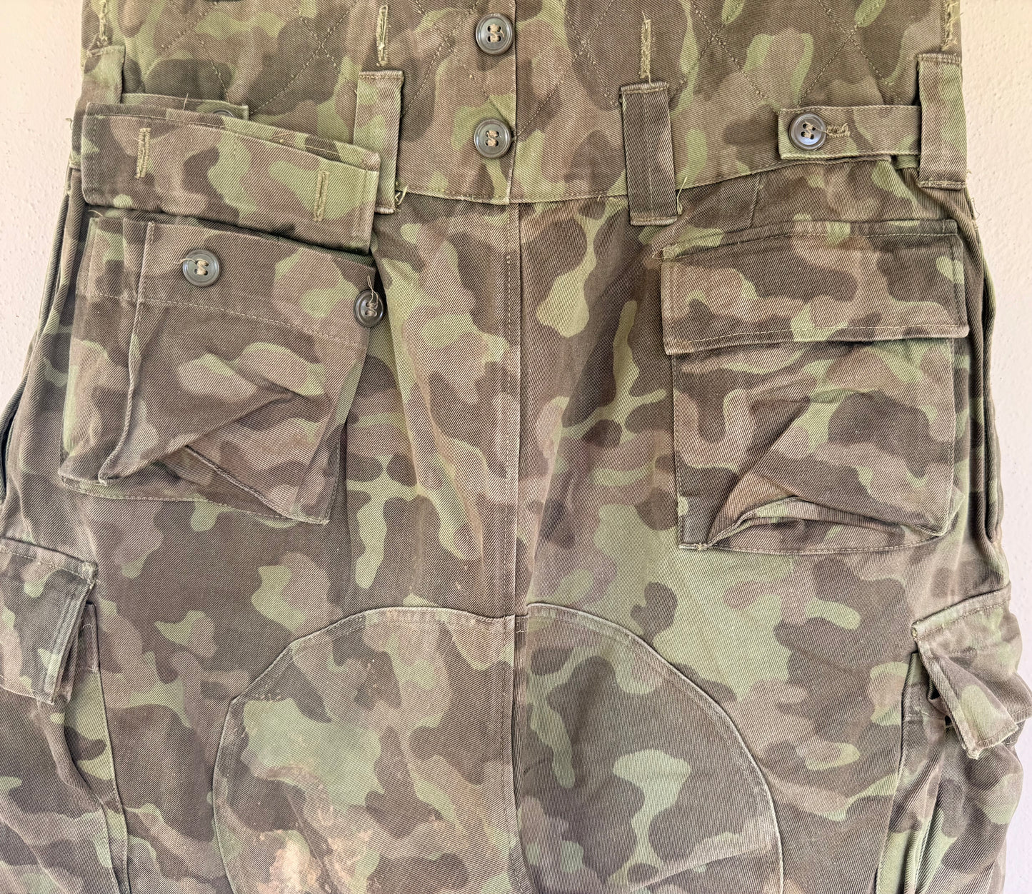 Close-up of a camouflage military-style utility garment, showing front flap pockets, button details, and a green-brown pattern on a plain light background.