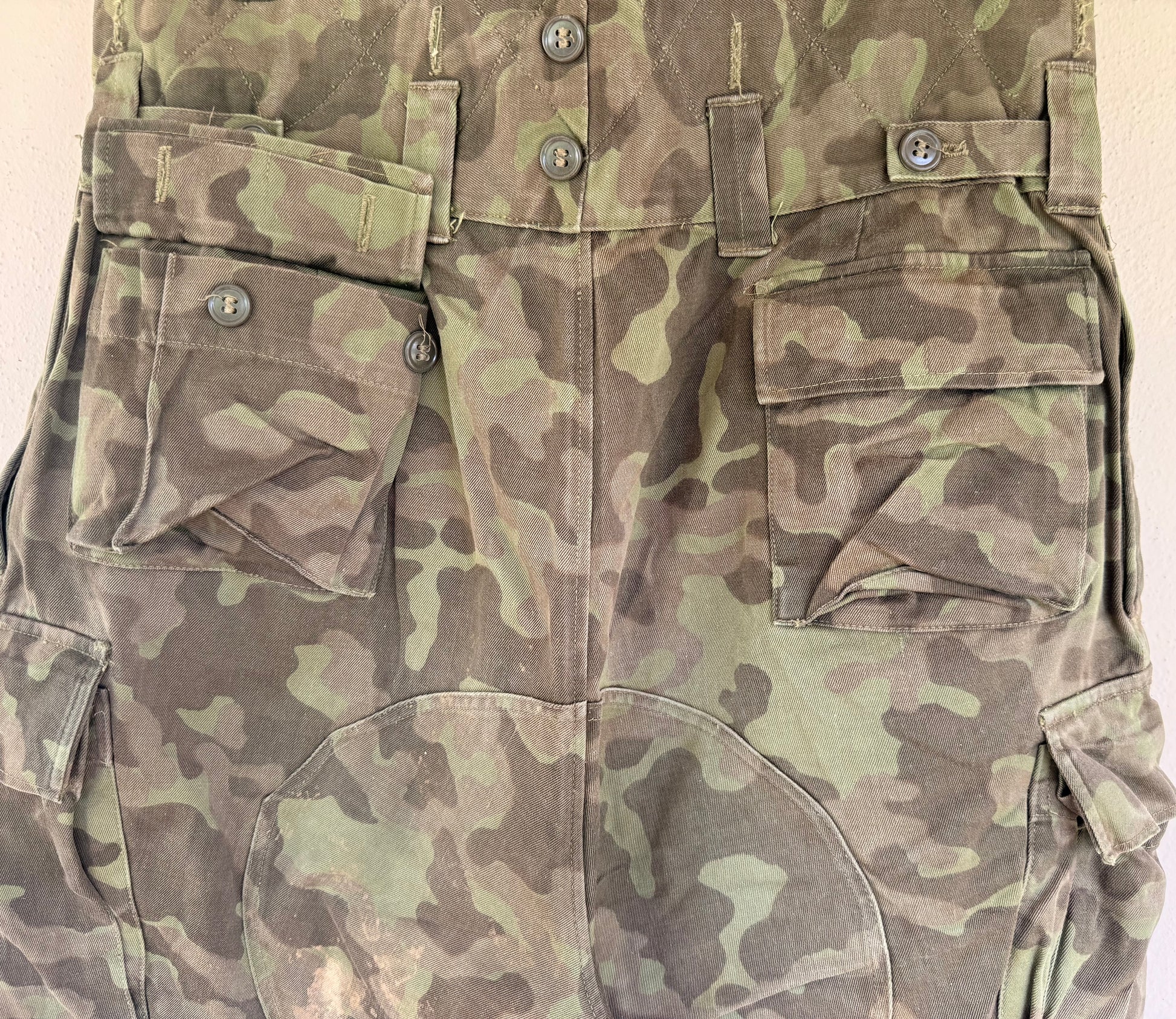 Close-up of a camouflage military-style utility garment, showing front flap pockets, button details, and a green-brown pattern on a plain light background.