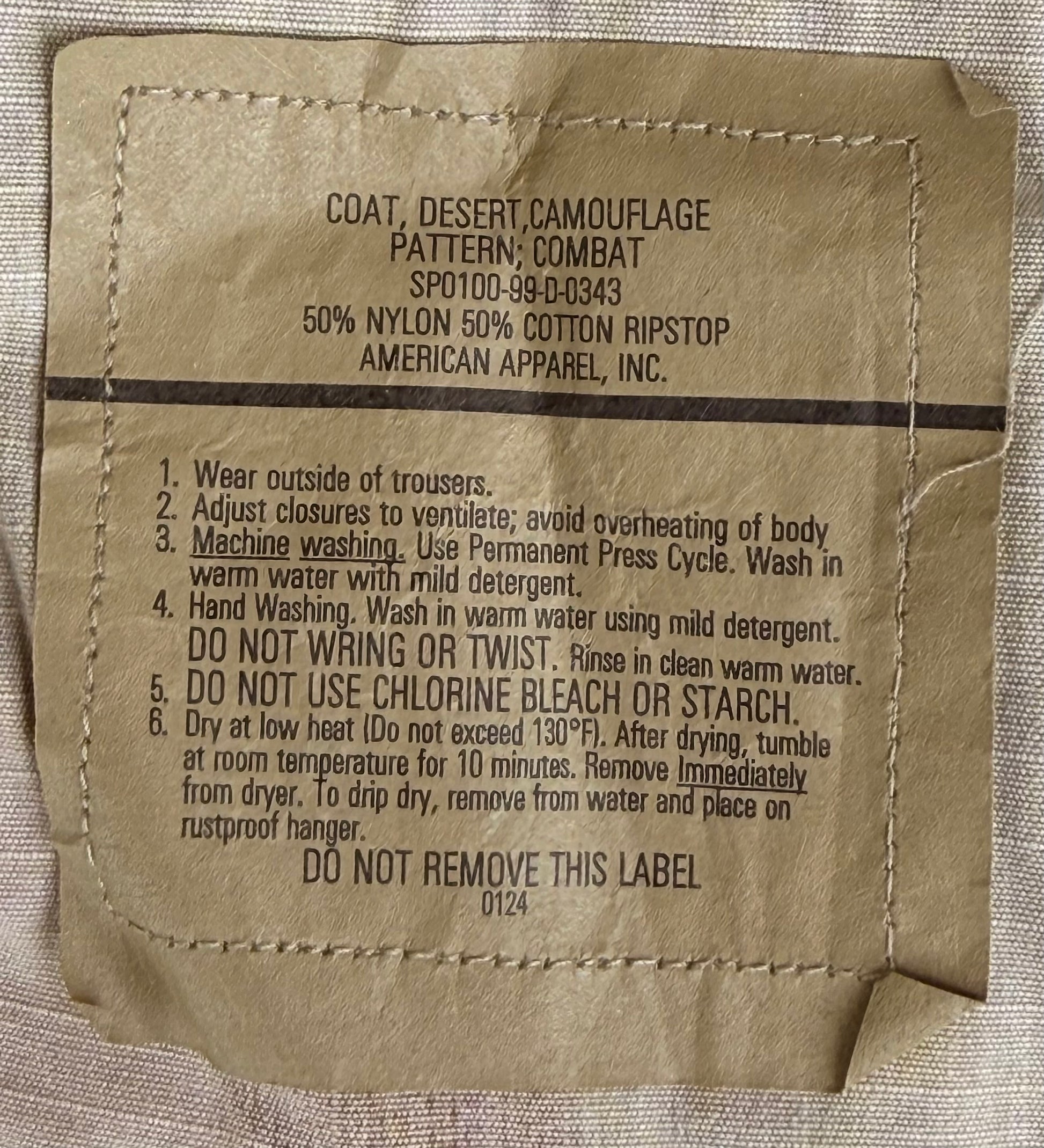 A close-up of a beige garment care label sewn onto light gray fabric. The label reads 'COAT, DESERT/CAMOUFLAGE PATTERN: COMBAT' and includes washing and care instructions, with 'AMERICAN APPAREL, INC.