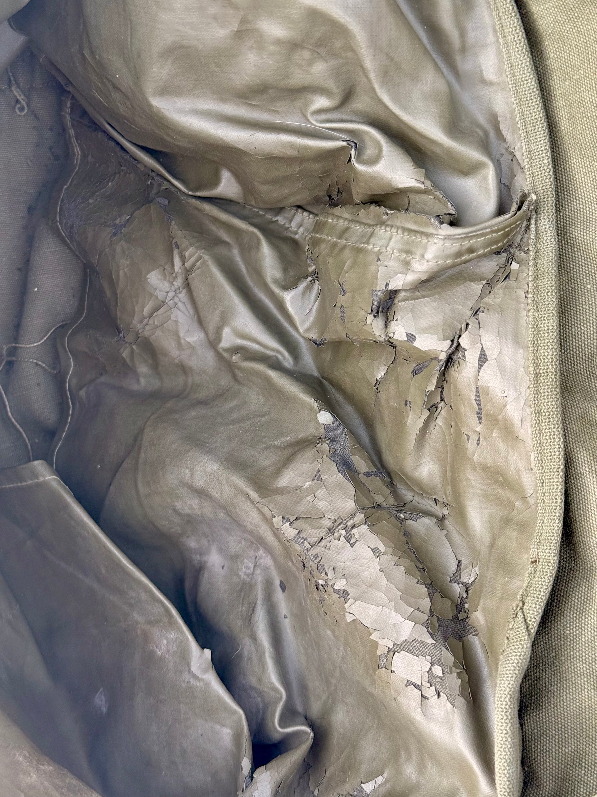 Close-up of a damaged olive green fabric item, showing a glossy, wrinkled interior lining with significant cracks and peeling layers exposing off-white material underneath.