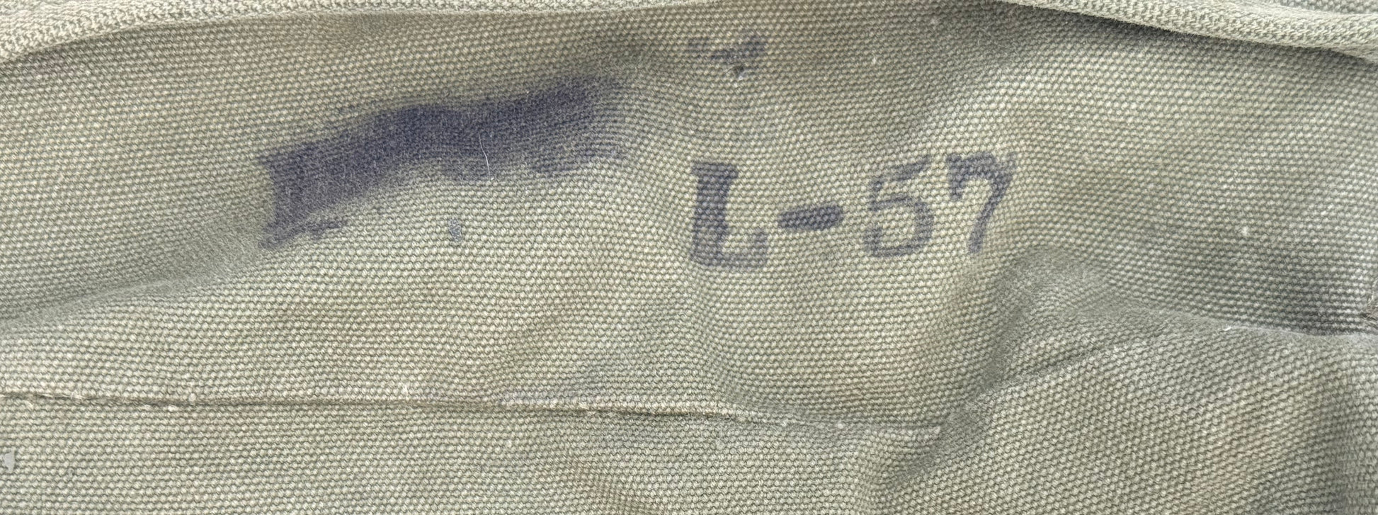 Close-up of a light olive drab canvas or nylon fabric surface with a dark blue-gray stamped marking reading 'L-57'.