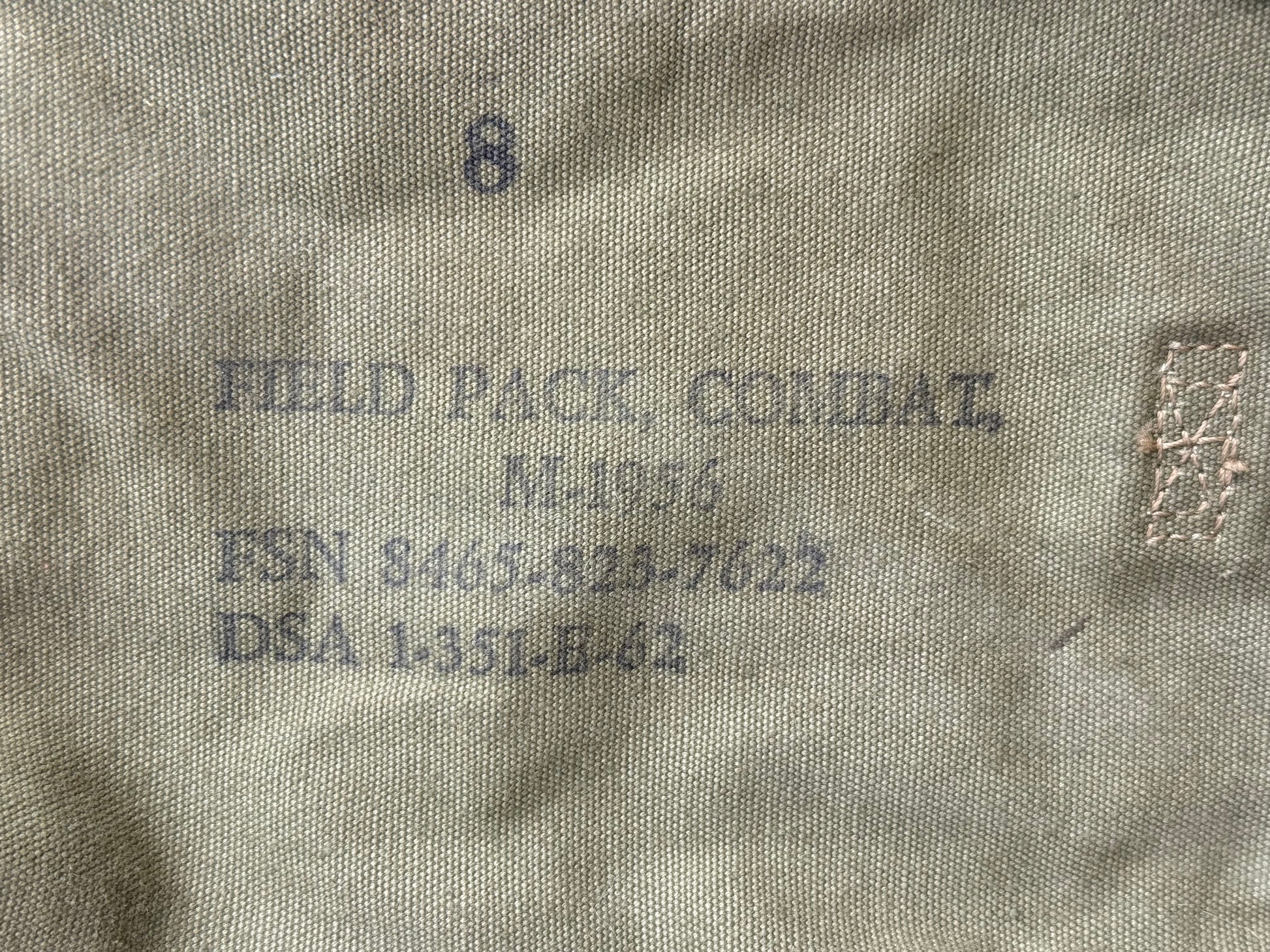 Close-up of a beige or khaki fabric surface with printed dark gray text reading 'FIELD PACK, COMBAT,' along with a number 8 and a small rectangular woven label or patch on the right.