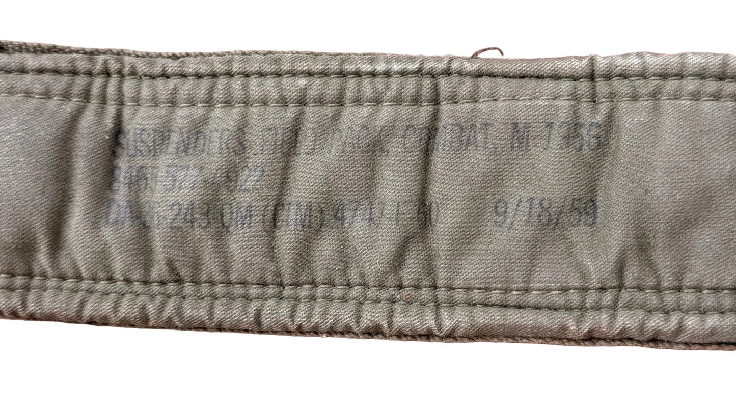 A close-up of a khaki olive green fabric strap with printed black text reading 'SUSPENDERS, FIELD PACK, COMBAT, M-795' and other serial numbers, laid flat on a textured pink background.