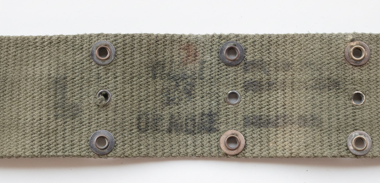 Close-up of a textured olive drab canvas strap with metal grommets, featuring faded US Navy markings, set against a pink background.