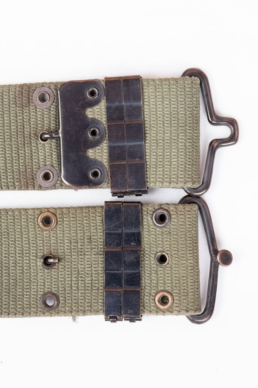 A close-up of a vintage watch strap with a woven khaki canvas, dark metal hardware, and a distinctive curved metal buckle, laid flat on a textured pink background.