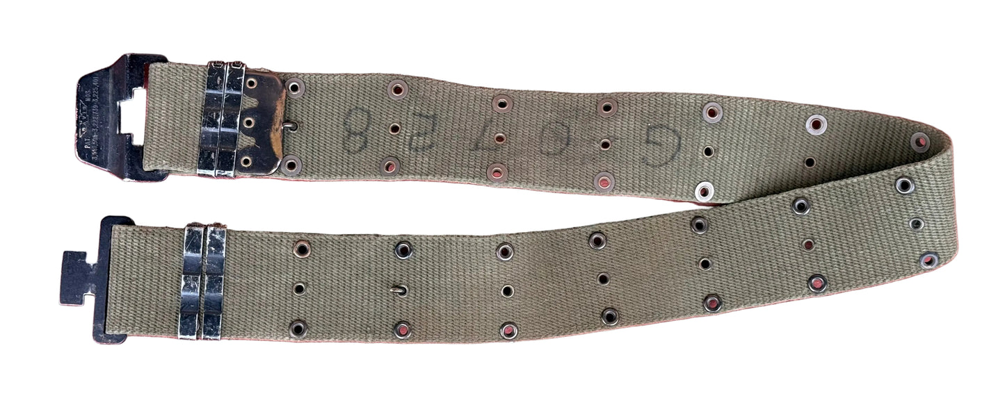 An olive drab canvas military-style belt with metal grommets and black plastic buckle hardware, laid flat on a textured red surface.