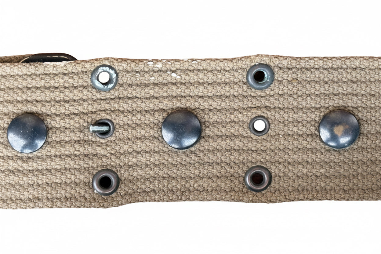 Close-up of a tan canvas strap with silver metal studs and grommets on a reddish-brown textured background.