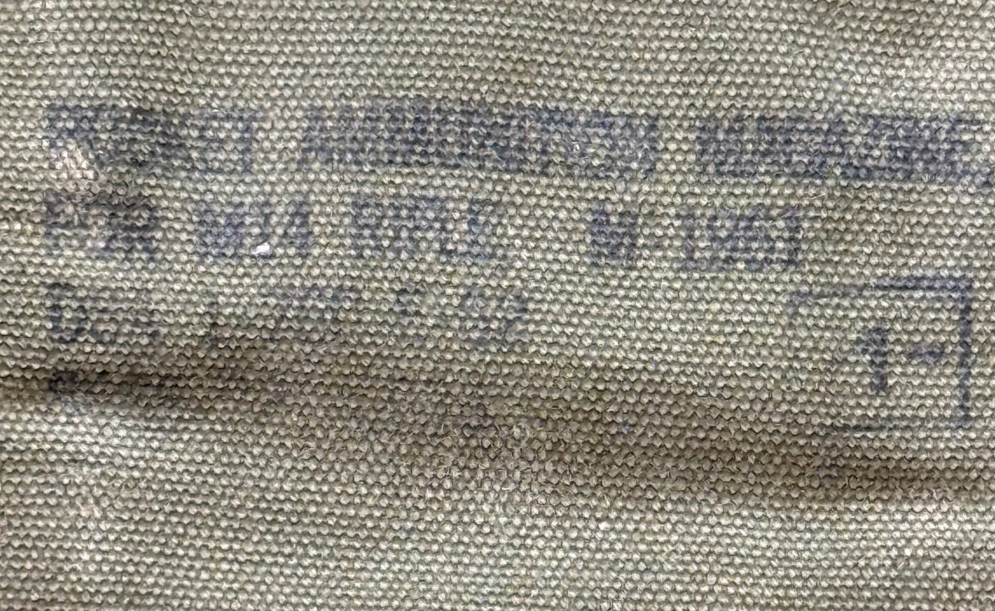Close-up of a textured beige canvas or woven fabric with faded blue printed text and a stamped box symbol.