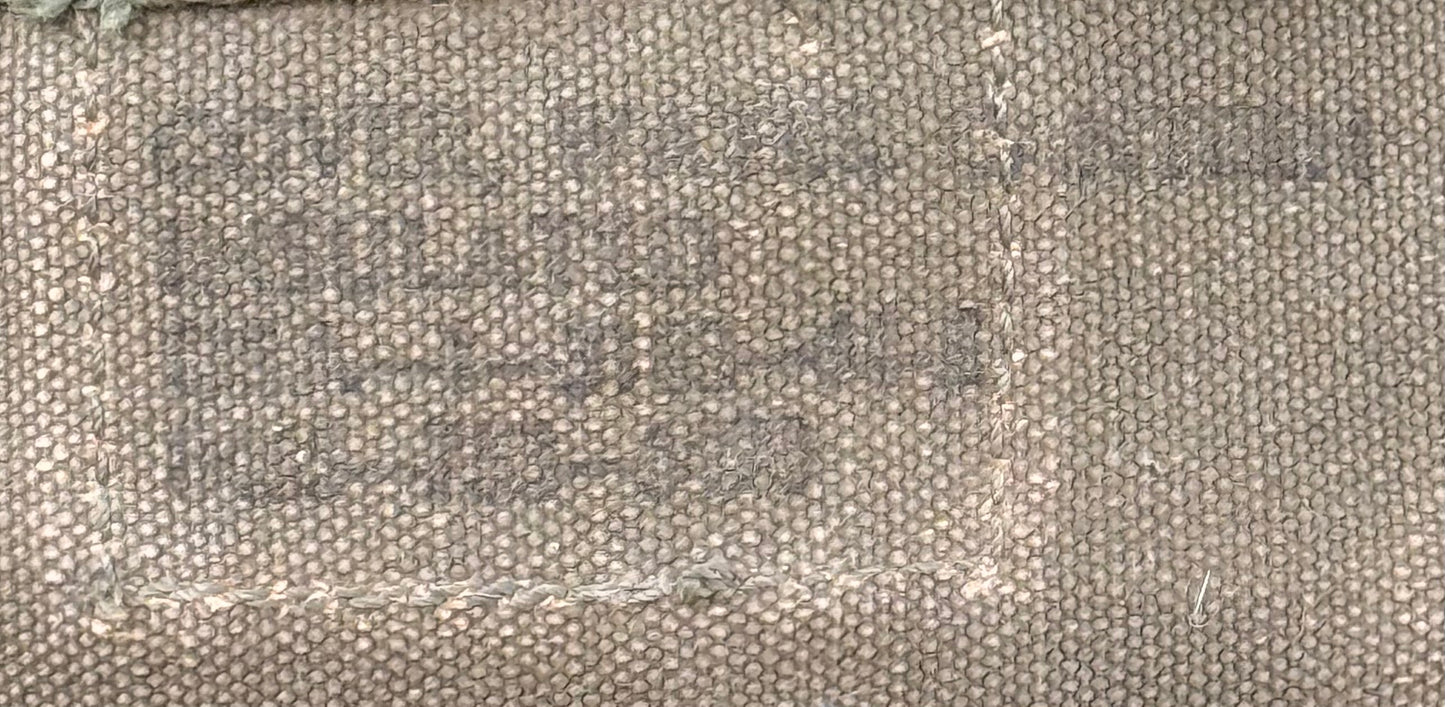 A close-up of a woven canvas or burlap material in a warm beige-gray color, showing a stitched rectangular patch area with faded stamped text.