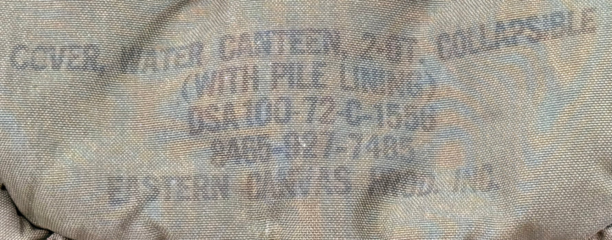 Close-up of a beige canvas cover with blue printed text reading 'COVER WATER CANTEEN 2 QT COLLAPSIBLE (WITH PILE LINING) OSA100-72-C-1596 9406-027-7746 EASTERN CANVAS PROD. INC.