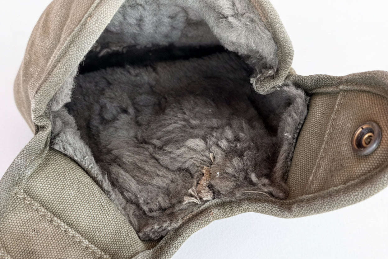 Close-up of the interior of a canvas pouch or bag, showing a gray faux-fur lining and an olive drab canvas exterior with a metal snap closure, set against a pink background.