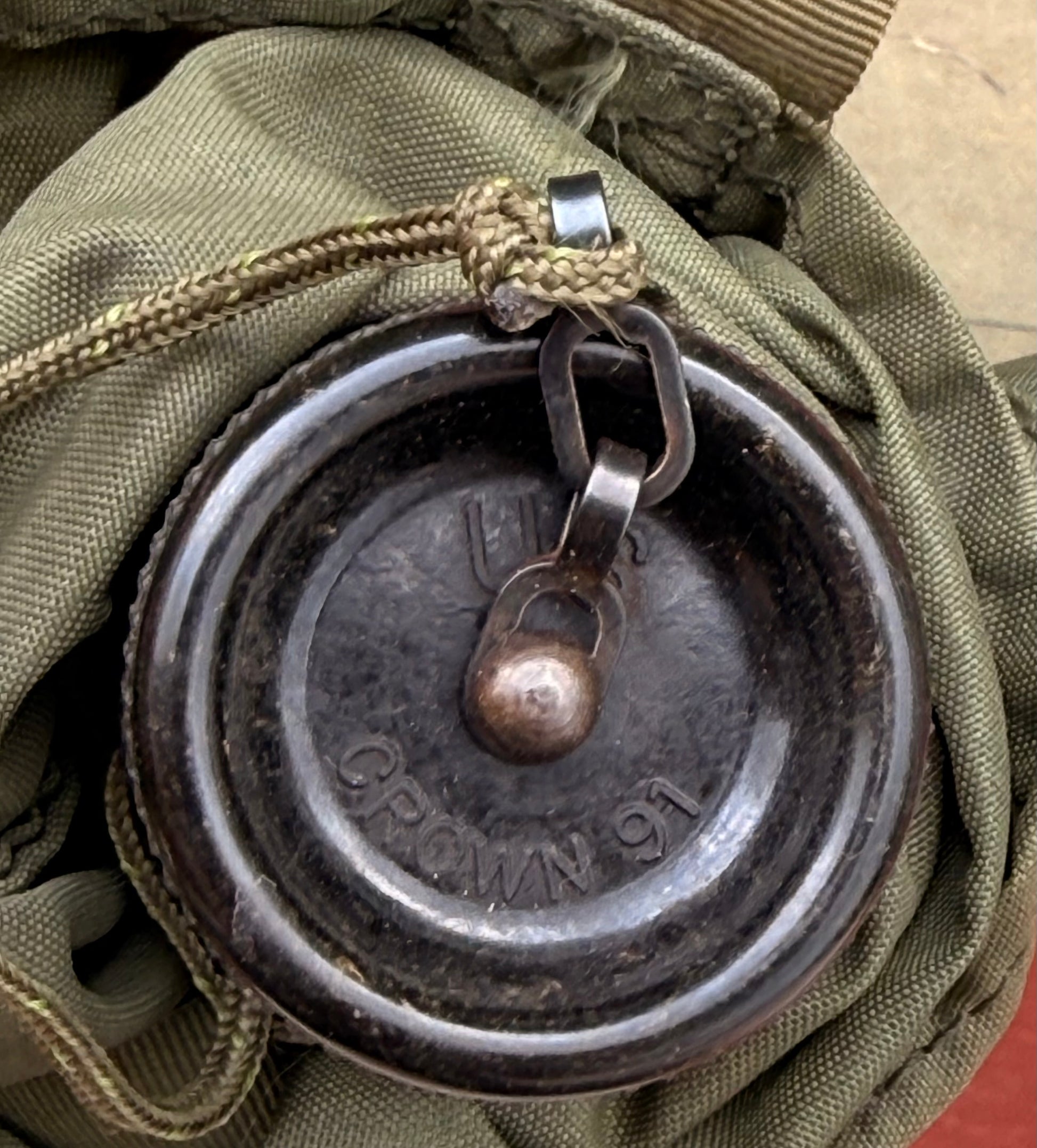 A close-up of a round metal lock or cap with a small padlock attached, resting on olive green canvas fabric.