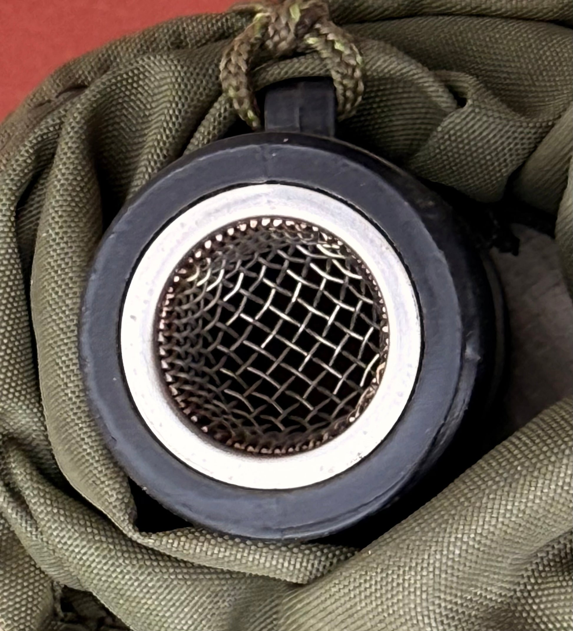 Close-up of a round black and silver metal mesh filter or valve component resting on olive green fabric.