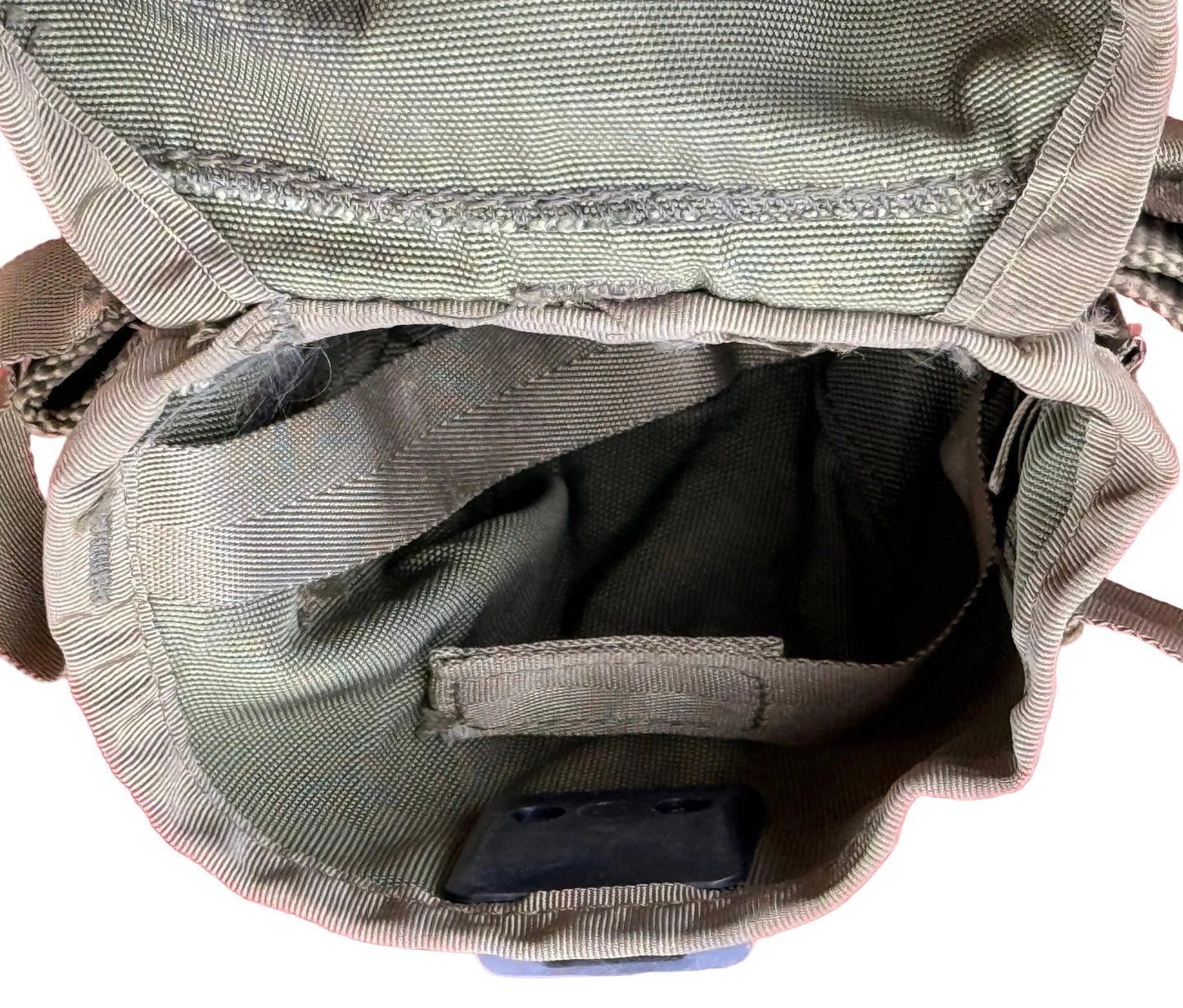 Close-up of the interior of a canvas pouch or small bag in olive drab green fabric, showing the inside lining, zipper area, and a small dark rectangular item resting inside, set against a pink textured background.