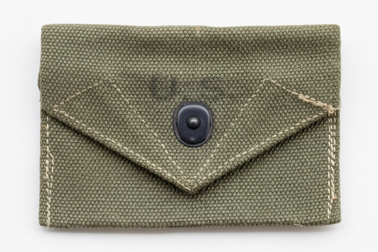 An olive green canvas envelope-style pouch with a black snap button closure, laid flat against a textured pink background.