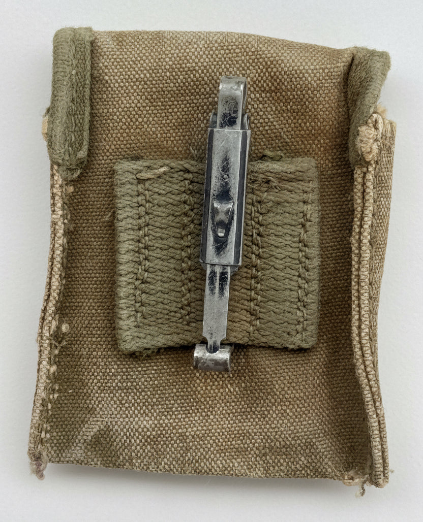 A vintage military-style canvas pouch in olive drab, featuring a central stitched pocket and a silver metal clip or fastener, shown against a textured red background.