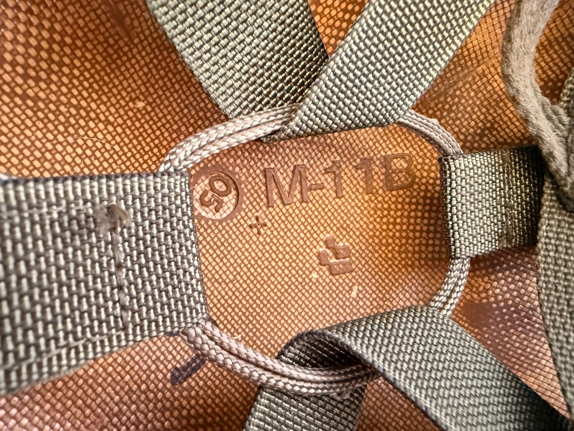 Close-up detail of a brown woven strap system with gray webbing and a central tan leather patch stamped with 'M-11B' and a logo.
