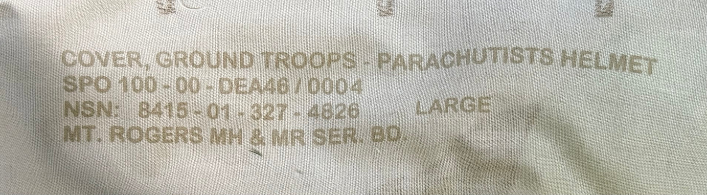 Close-up of a beige military fabric label with printed text reading 'COVER, GROUND TROOPS - PARACHUTISTS HELMET' and other serial information.