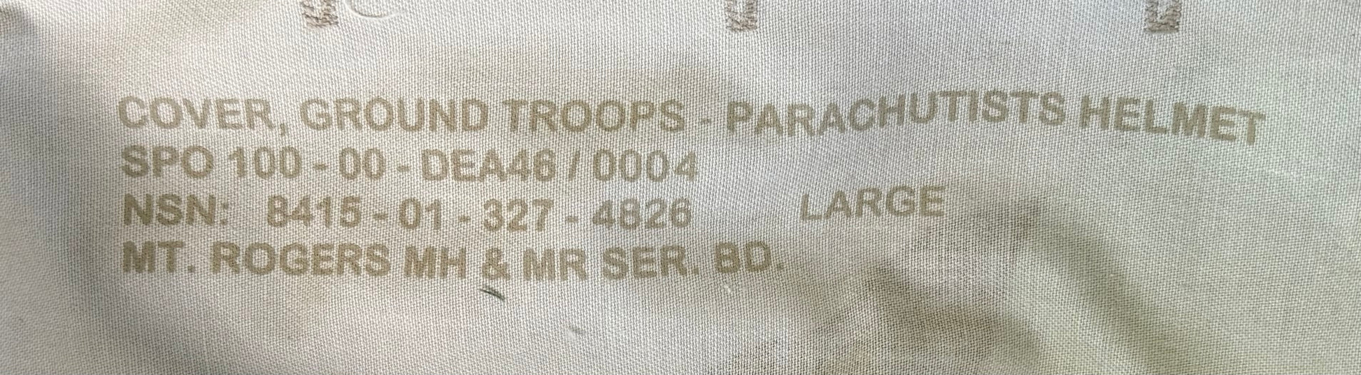 Close-up of a beige military fabric label with printed text reading 'COVER, GROUND TROOPS - PARACHUTISTS HELMET' and other serial information.
