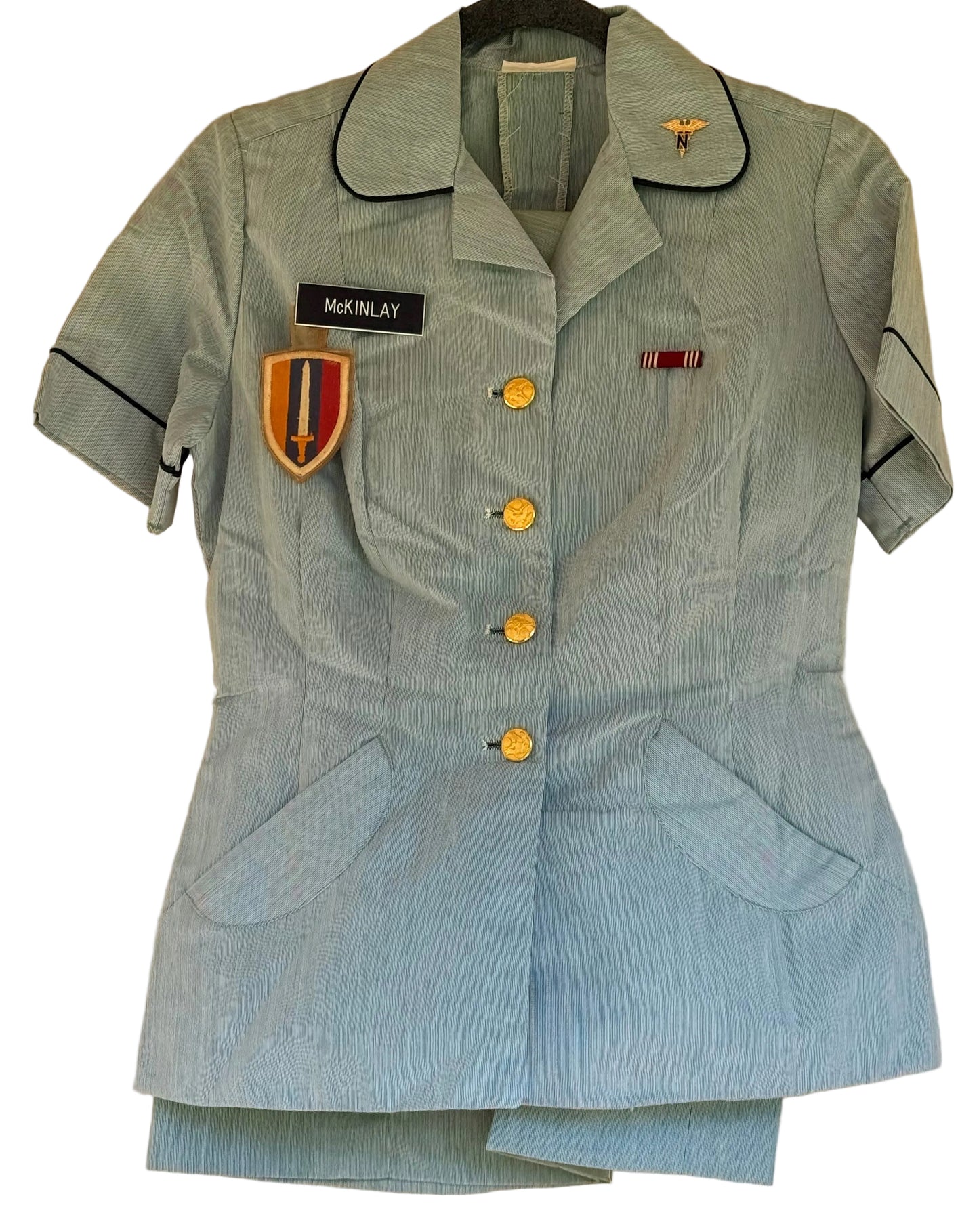 A light blue military-style nurse uniform jacket with short sleeves, gold buttons, black piping, and embroidered insignia, hanging against a beige wall.