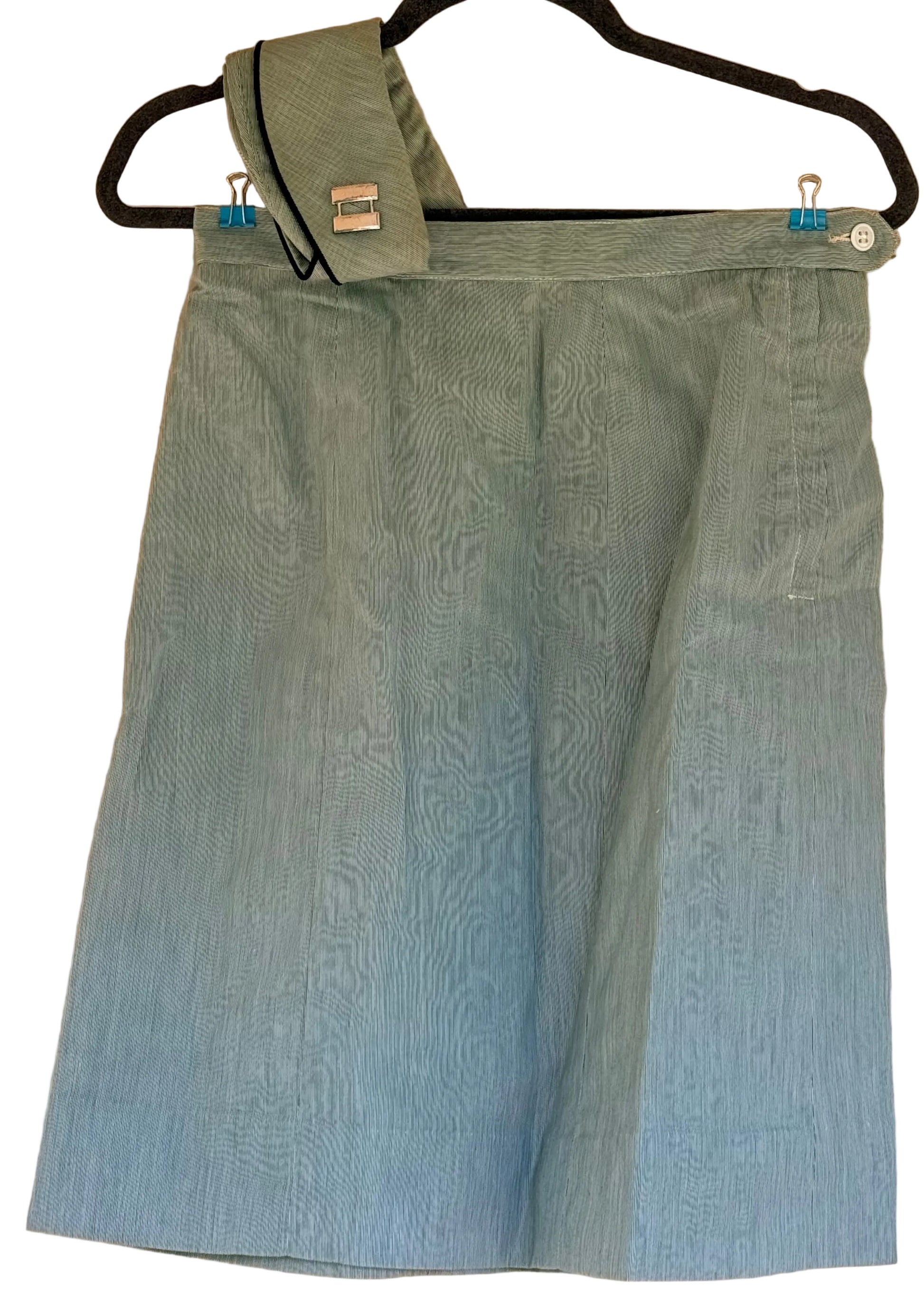 A light blue textured skirt with a matching fabric belt and a small silver tag, hanging on a black hanger against a beige wall.