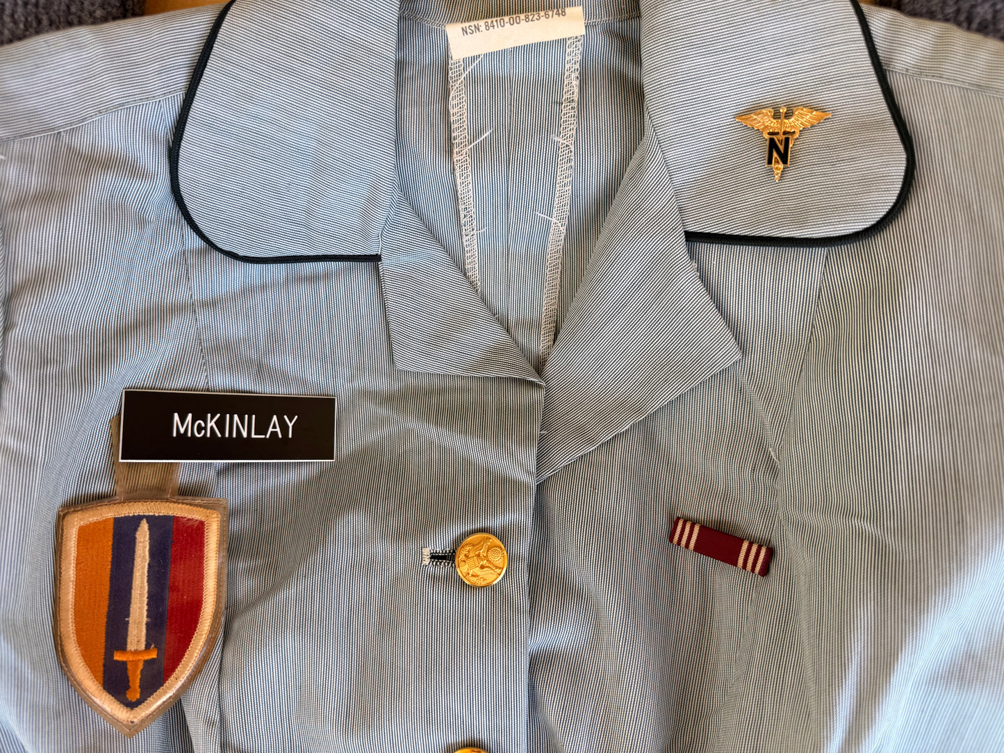 A light blue military-style uniform jacket with black piping, featuring a gold medical insignia on the collar, a black name tape reading 'MCKINLAY', a shield-shaped embroidered patch with a sword, and a small ribbon badge.
