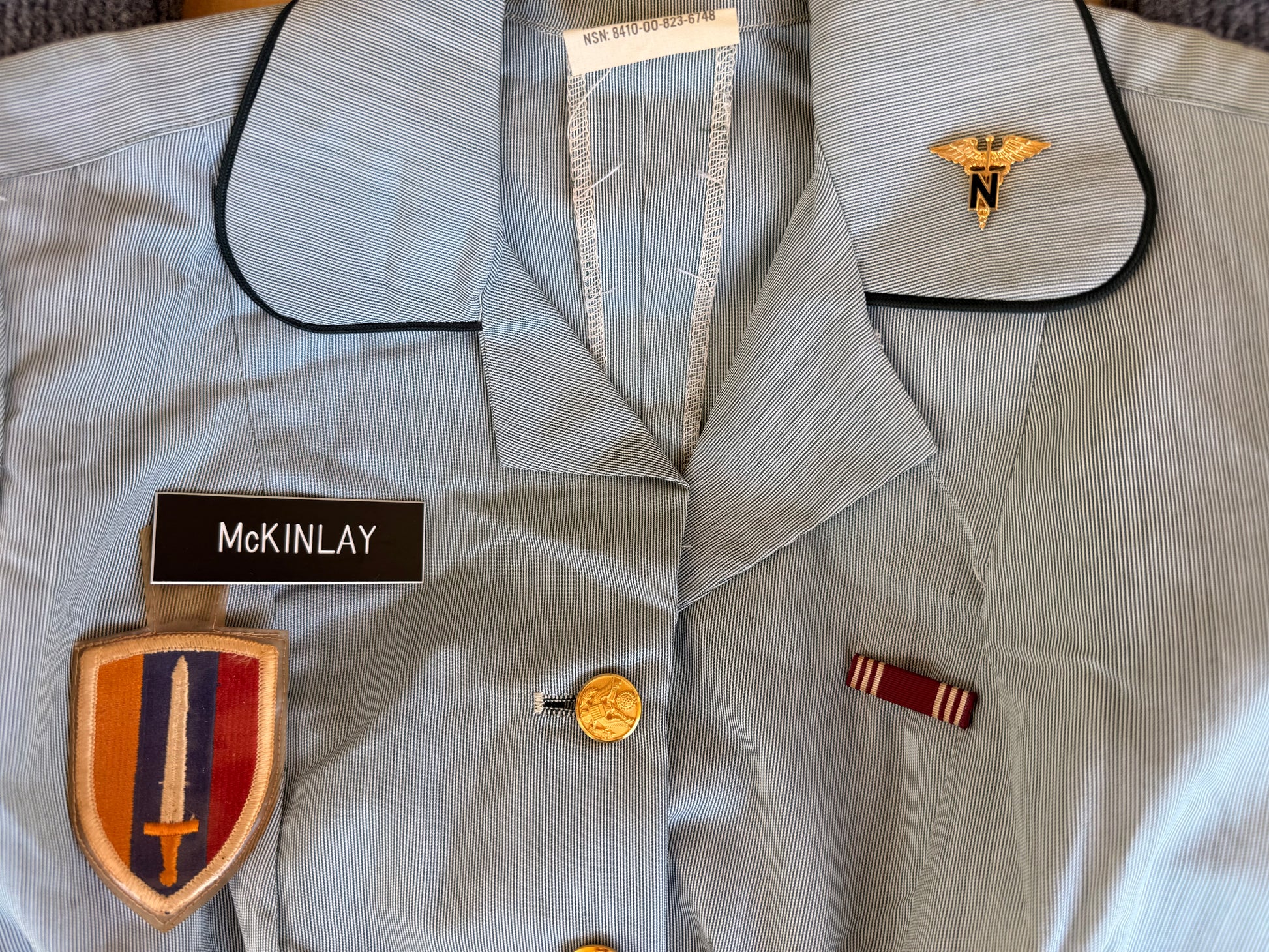 A light blue military-style uniform jacket with black piping, featuring a gold medical insignia on the collar, a black name tape reading 'MCKINLAY', a shield-shaped embroidered patch with a sword, and a small ribbon badge.