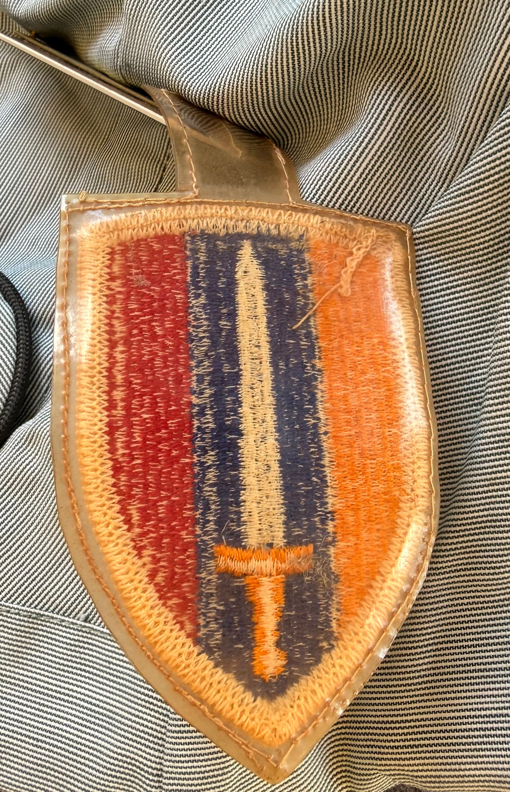 A shield-shaped embroidered patch with a tan leather border, featuring a central vertical tricolor design of red, navy blue, and orange with a gold sword motif, laid flat on a gray and white striped fabric background.