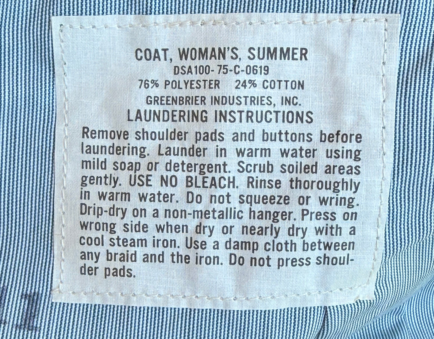 Close-up of a white rectangular laundry instruction label sewn onto a blue and white striped women's summer coat. The label text includes the brand name Greenbrier Industries, Inc.