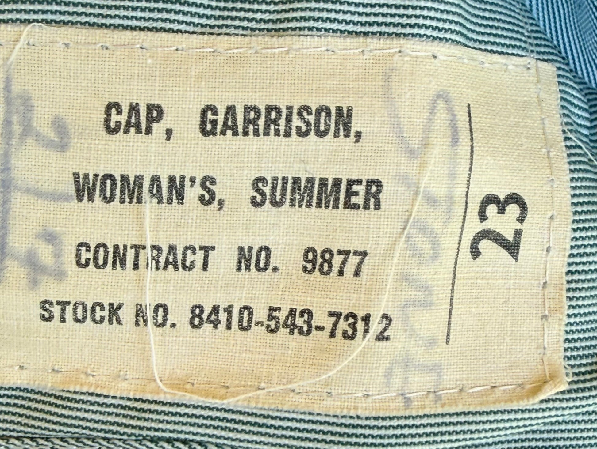 Close-up of a sewn fabric label reading 'CAP, GARRISON, WOMAN'S, SUMMER CONTRACT NO. 9877 STOCK NO.