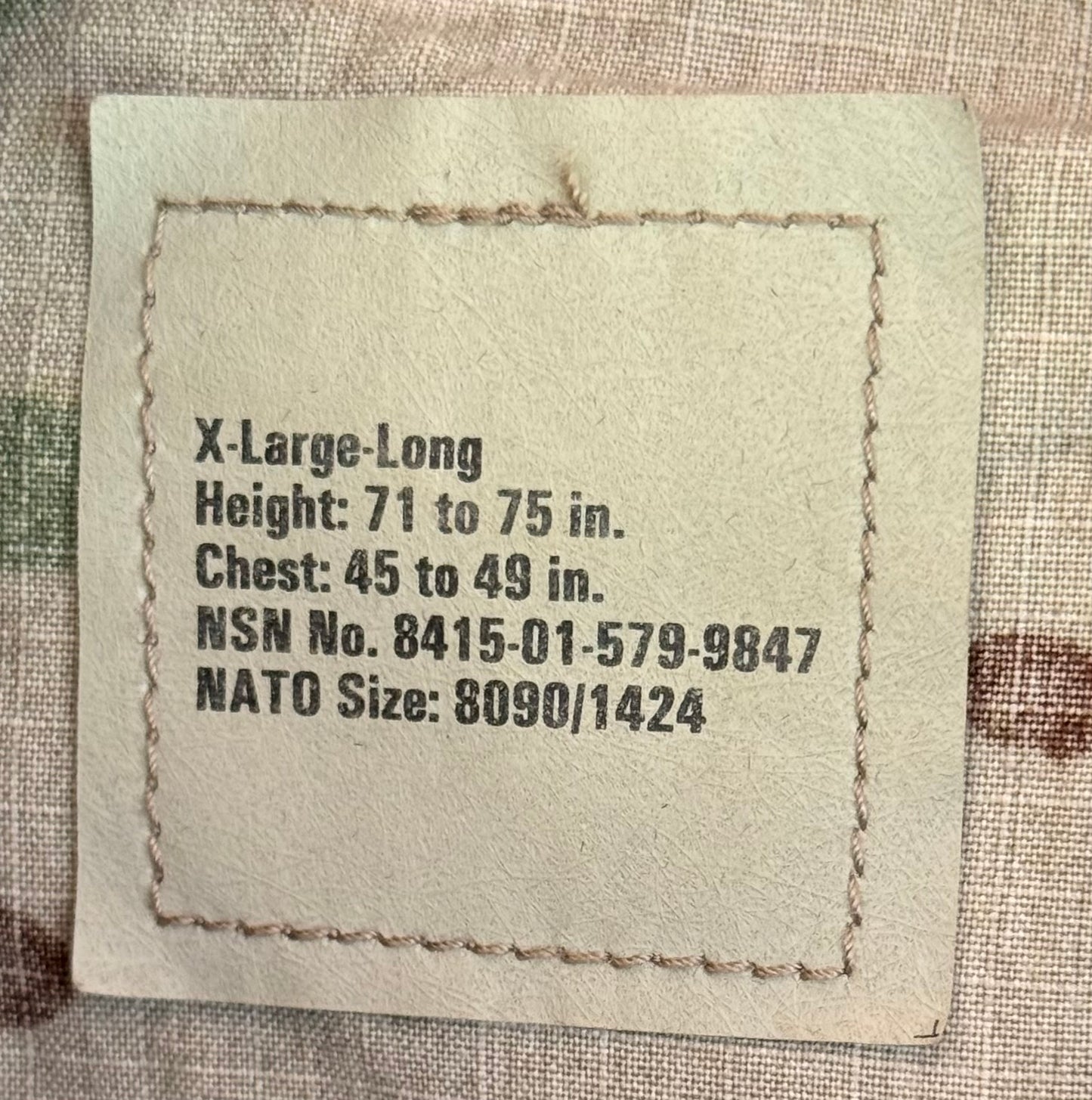 A square beige fabric label with black printed sizing information for X-Large-Long, including height, chest, NSN number, and NATO size, placed on a textured woven fabric background.
