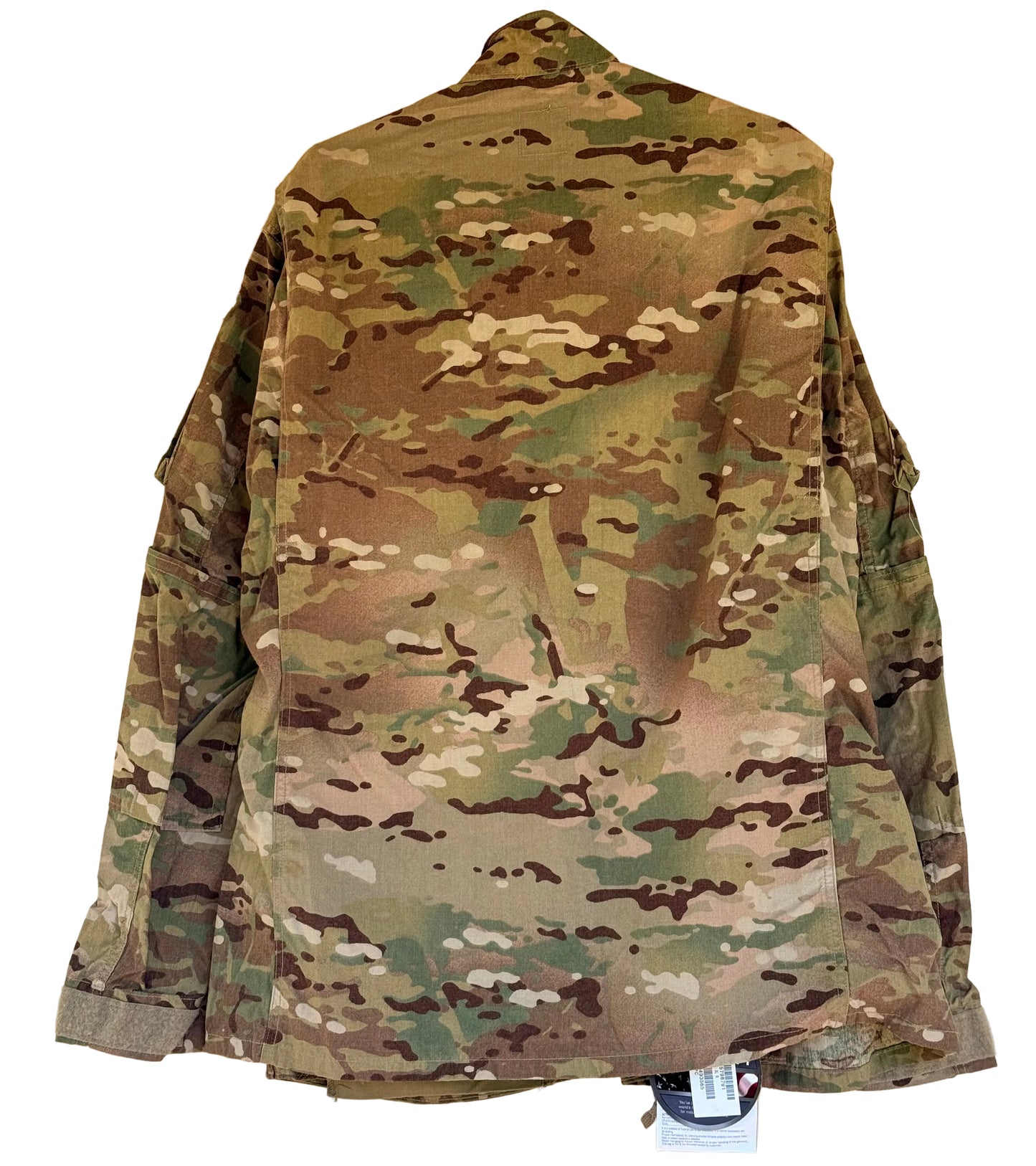 A military-style camouflage jacket shown from the back, hanging against a plain beige wall. The garment features a multi-terrain pattern with shades of green, brown, tan, and black, and has a small retail tag attached at the bottom hem.