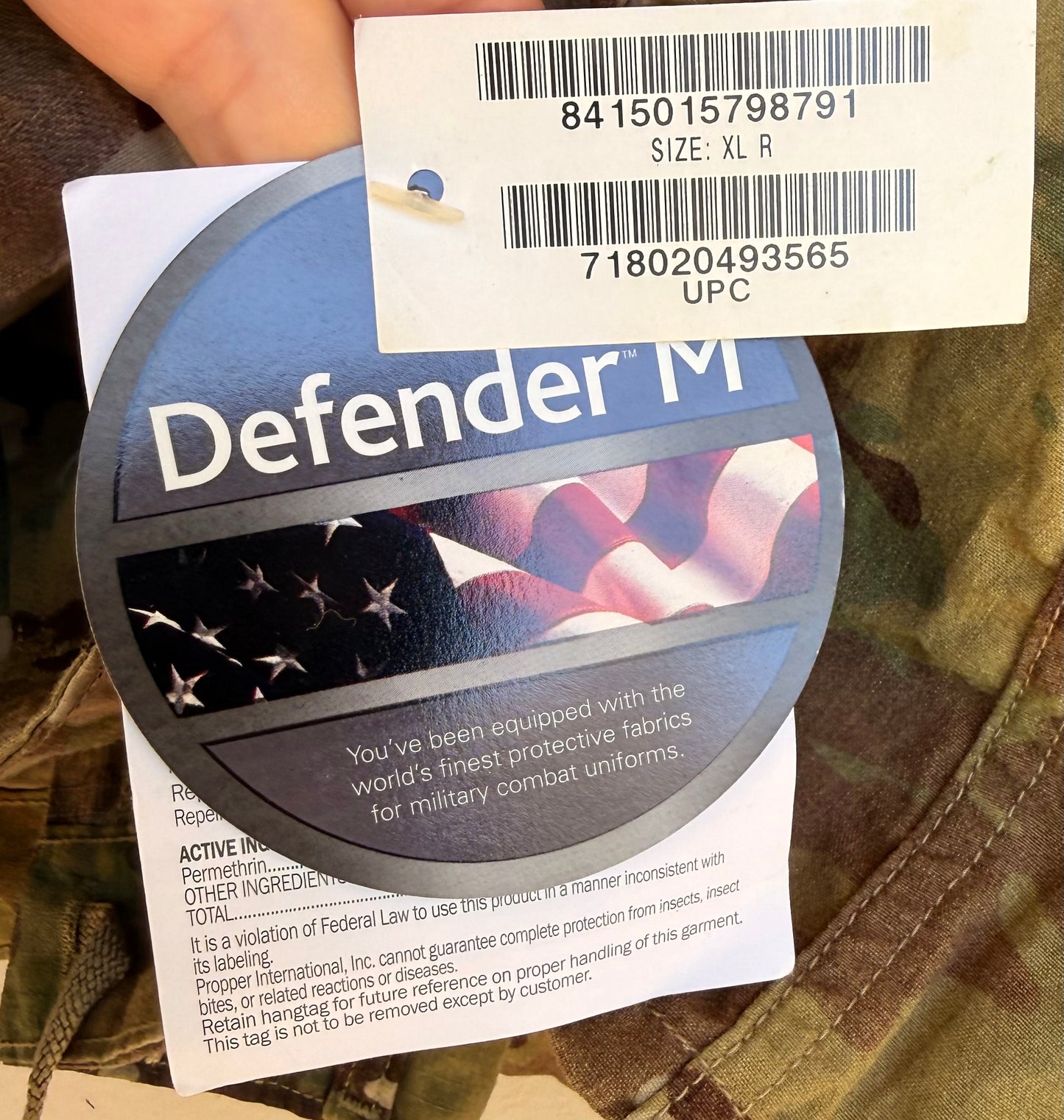 A close-up of a circular Defender M military fabric tag attached to a white barcode sticker, showing the text 'Defender M' and fabric information, with a hand holding camouflage clothing in the background.