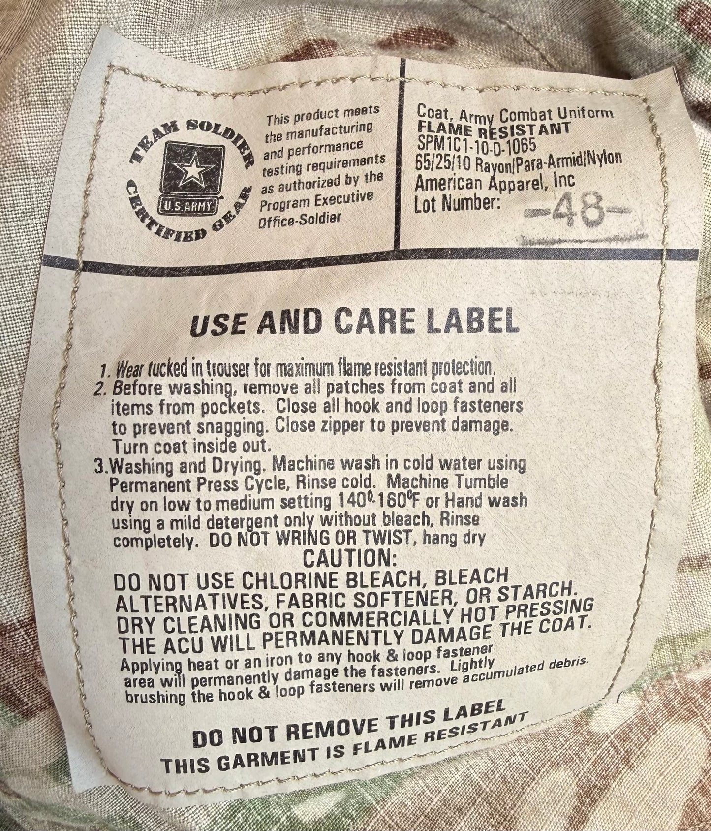 A close-up of a vintage military garment care label sewn onto a beige fabric tag. The label features the 'Team Soldier Certified Gear' logo and provides instructions for washing and care, including specifying a medium-low setting on the machine and avoiding chlorine bleach, fabric softener, or starch.
