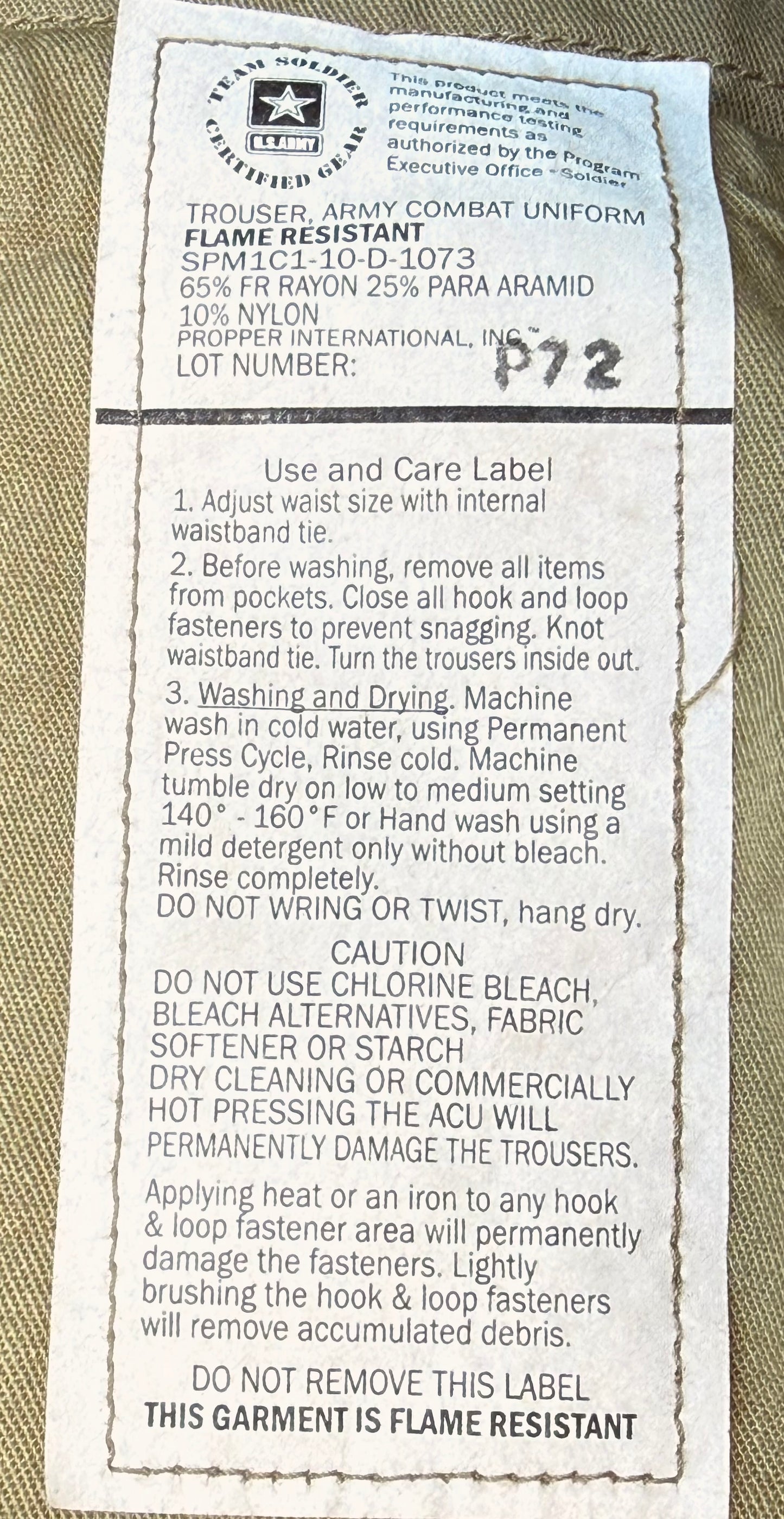 A close-up of a beige military-style uniform fabric with a sewn-on care and size label. The label reads 'Trouser Army Combat Uniform' and lists material as 65% cotton 25% para aramid 10% nylon, with care instructions and a lot number 1972.