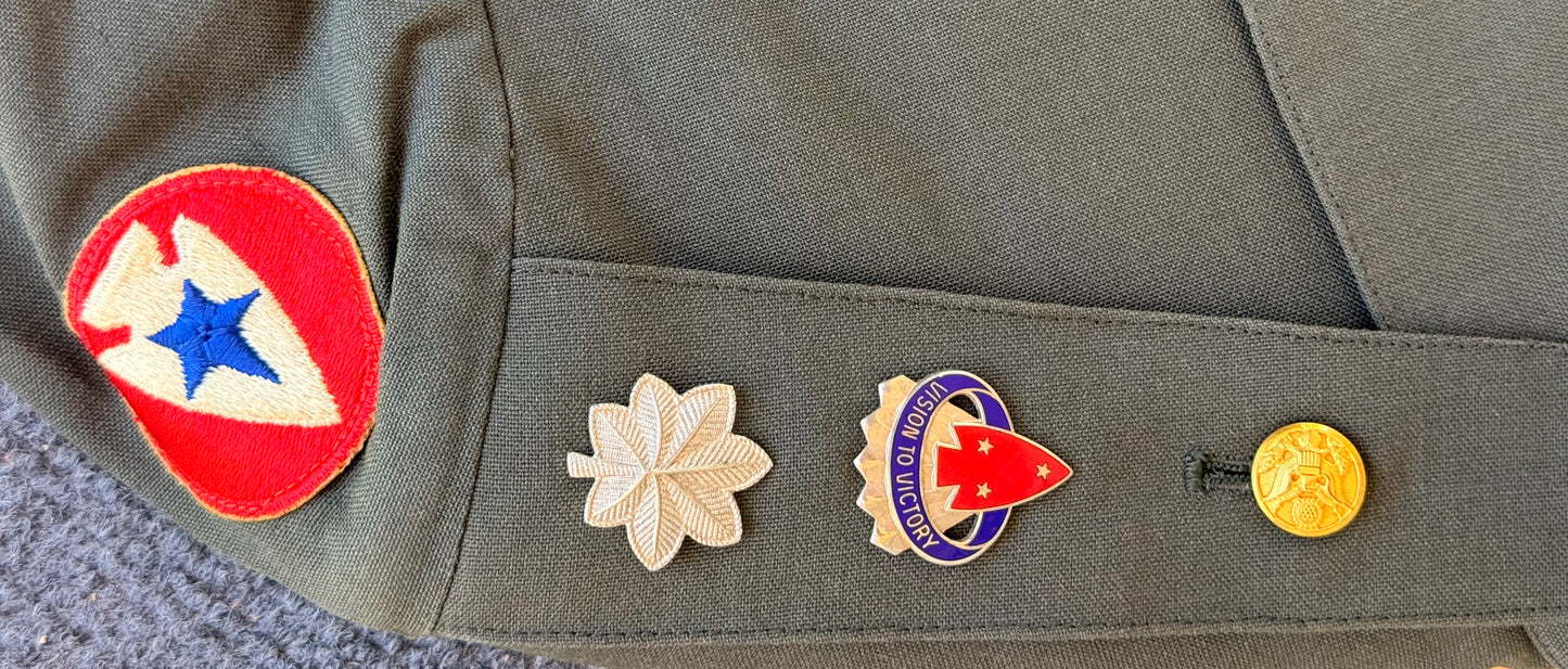 Close-up of an olive drab military uniform sleeve featuring a red, white, and blue embroidered patch on the upper sleeve and three embroidered insignia pins on the cuff pocket.