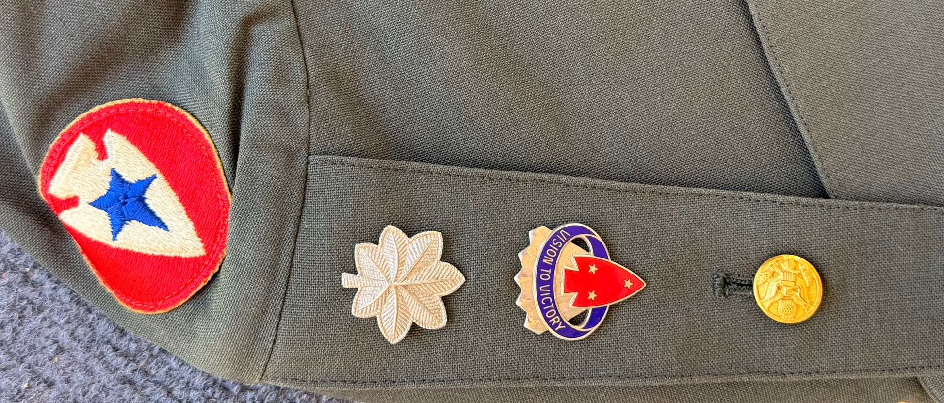 Close-up of an olive drab military uniform sleeve featuring a red, white, and blue embroidered patch on the upper sleeve and three embroidered insignia pins on the cuff pocket.