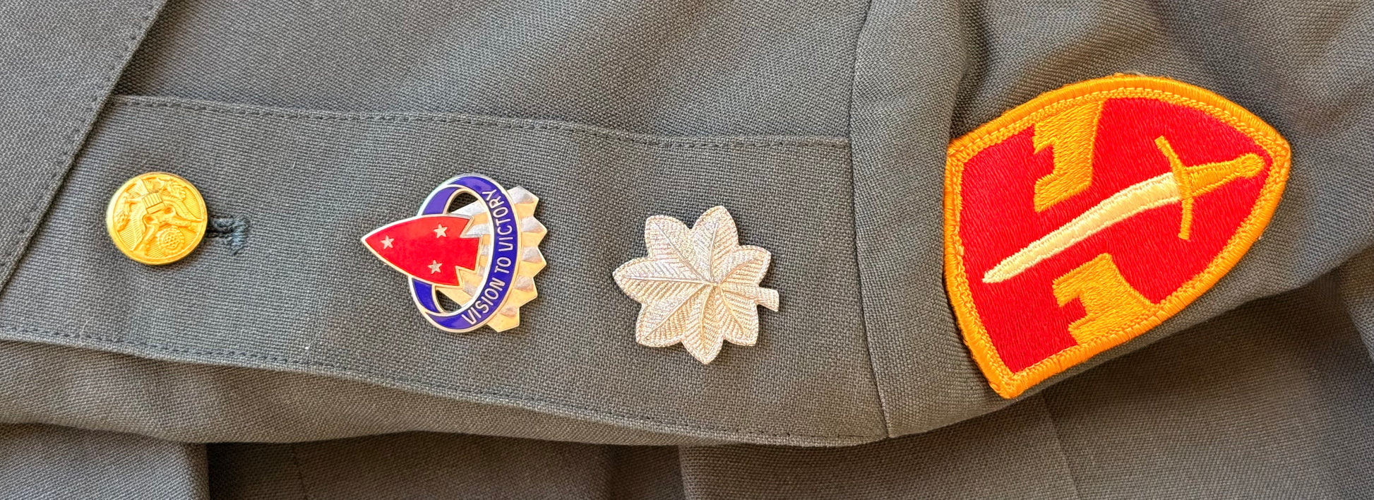 Close-up of a military uniform jacket showing three embroidered insignia pins on the left chest pocket: a round gold emblem, a red, white, and blue arrowhead insignia, and a white flower, alongside a red and yellow shield patch.