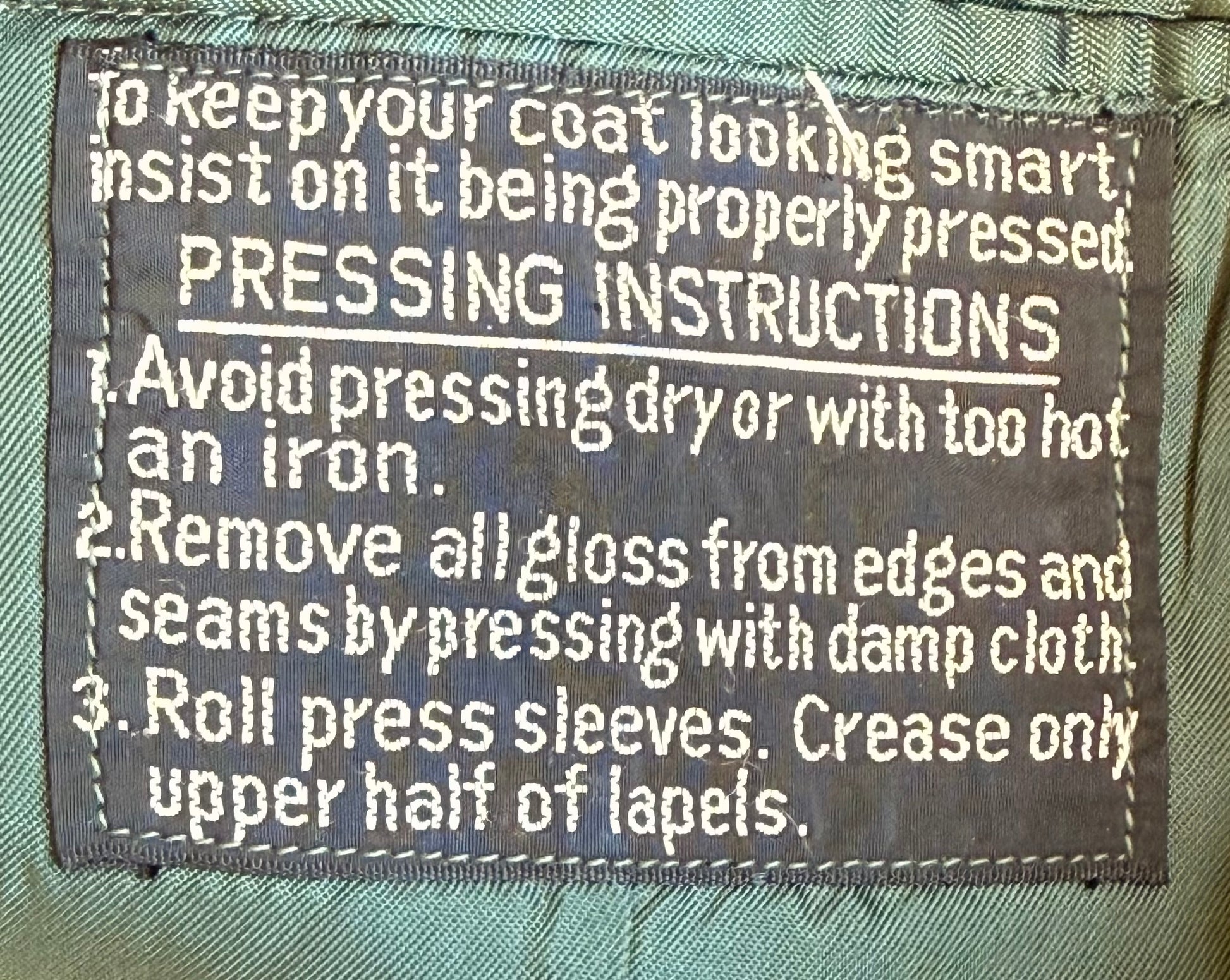 A close-up of a sewn fabric label with white text reading pressing instructions for a coat, including avoiding dry ironing and rolling press sleeves.