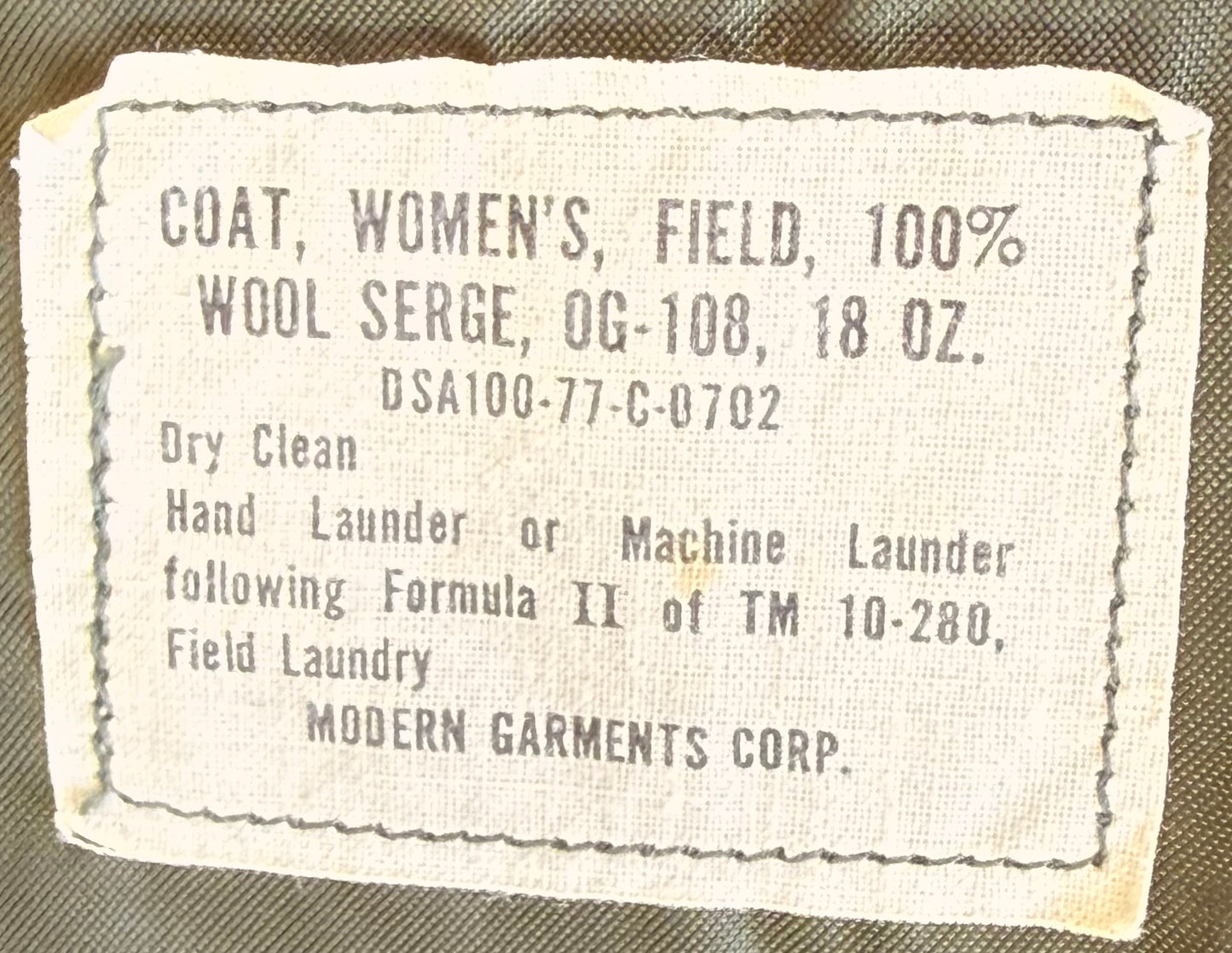 A close-up of a vintage fabric garment label reading 'COAT, WOMEN'S, FIELD, 100% WOOL SERGE, OG-108, 18 OZ.' with care instructions and 'MODERN GARMENTS CORP.