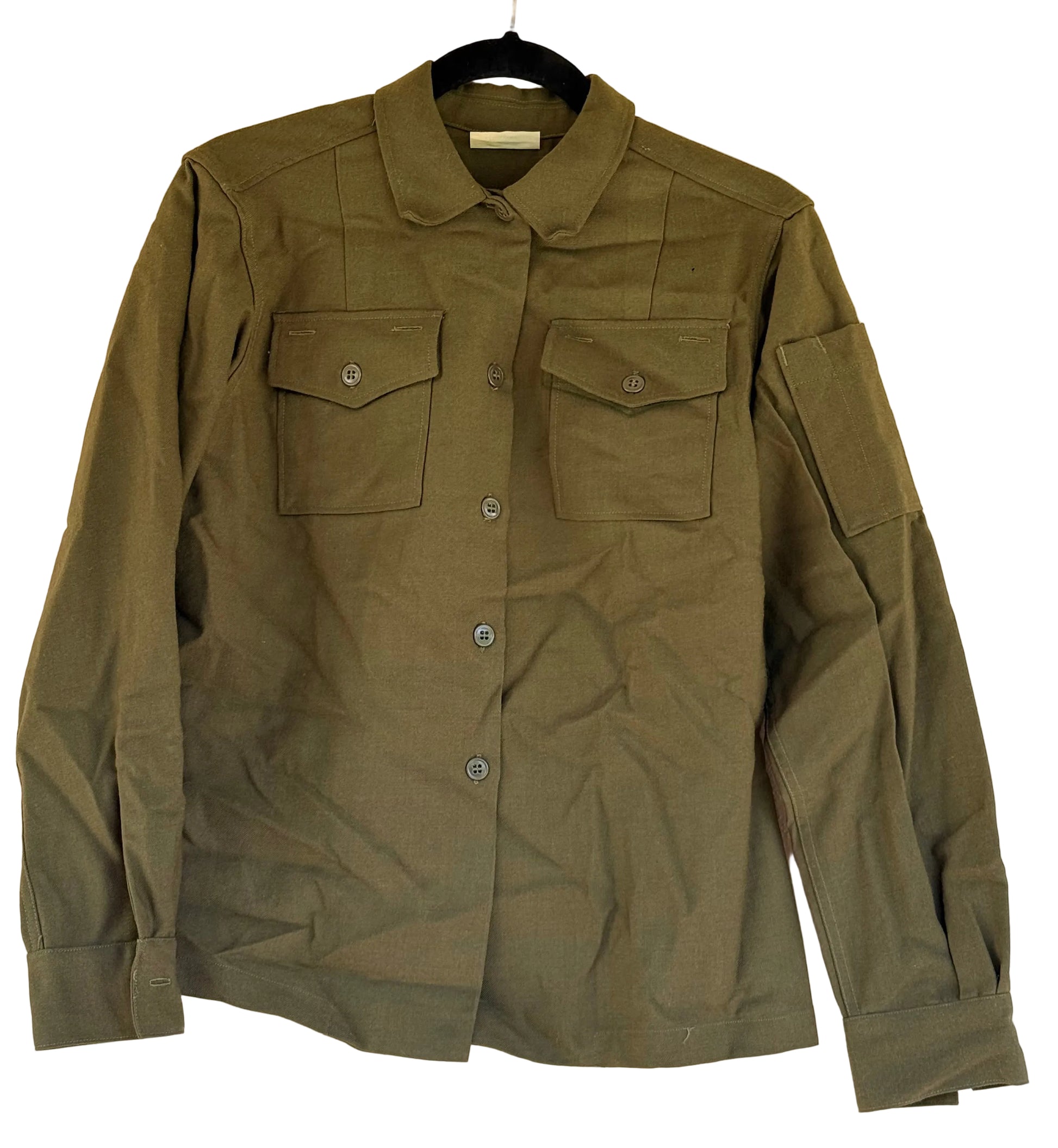 An olive green military-style button-up shirt with two chest flap pockets and a sleeve patch pocket, hanging on a black hanger against a textured beige wall.